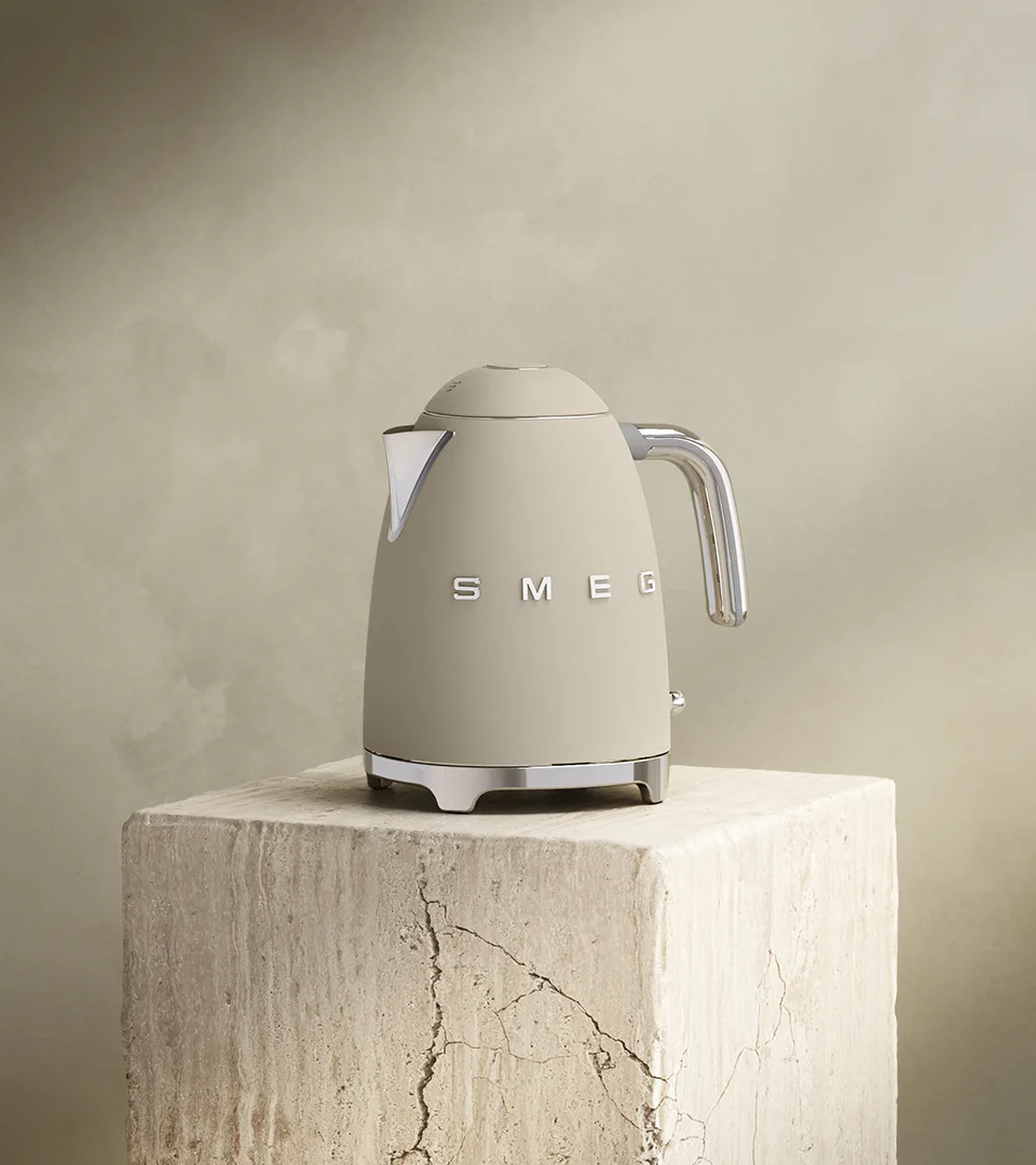 En matt beige Smeg vattenkokare med kromhandtag och bas står på ett texturerat beigt block mot en dämpad bakgrund.