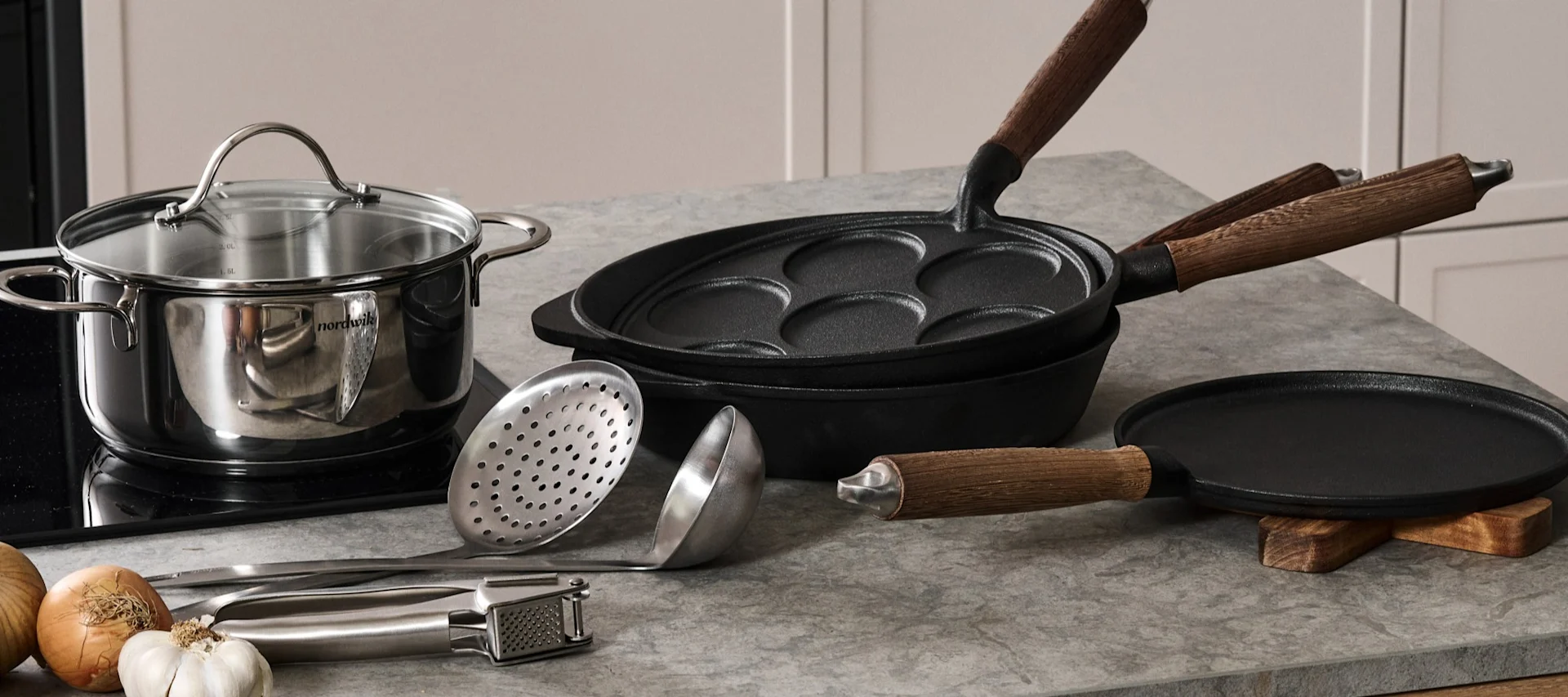 Kastrull i rostfritt stål, gjutjärnsplättlagg och stekpanna med diverse köksredskap och råvaror på en bänkskiva.