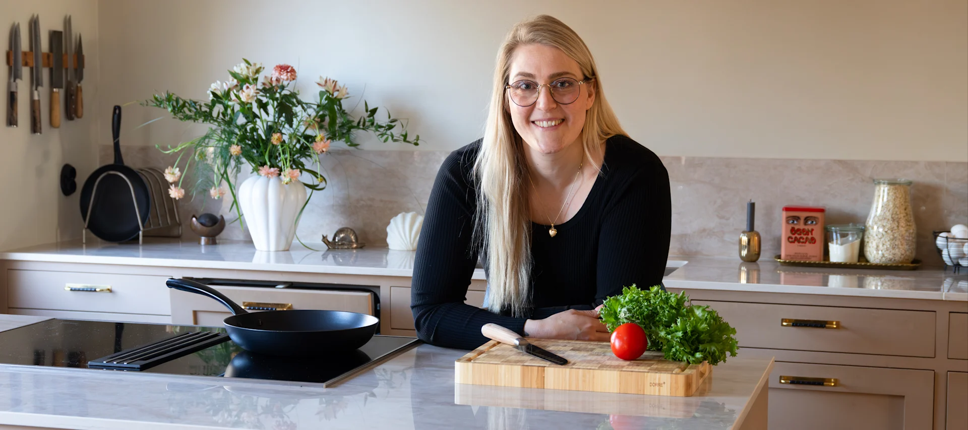 Leende kvinna i glasögon i modernt kök med panna på spisen, skärbräda med kniv, tomat och sallad.