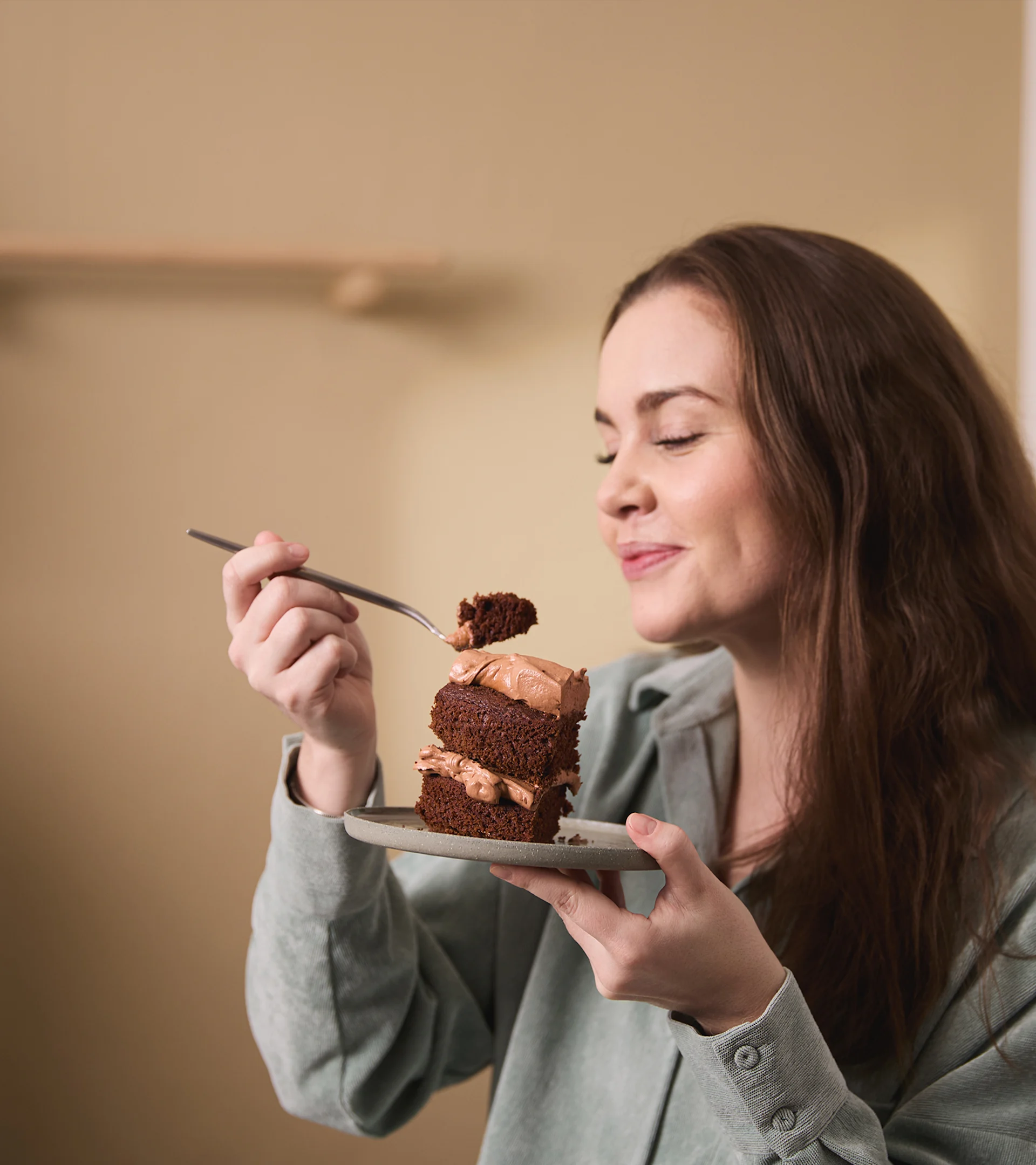 Kvinna med långt brunt hår, slutna ögon, njuter av en bit chokladkaka från en liten tallrik.