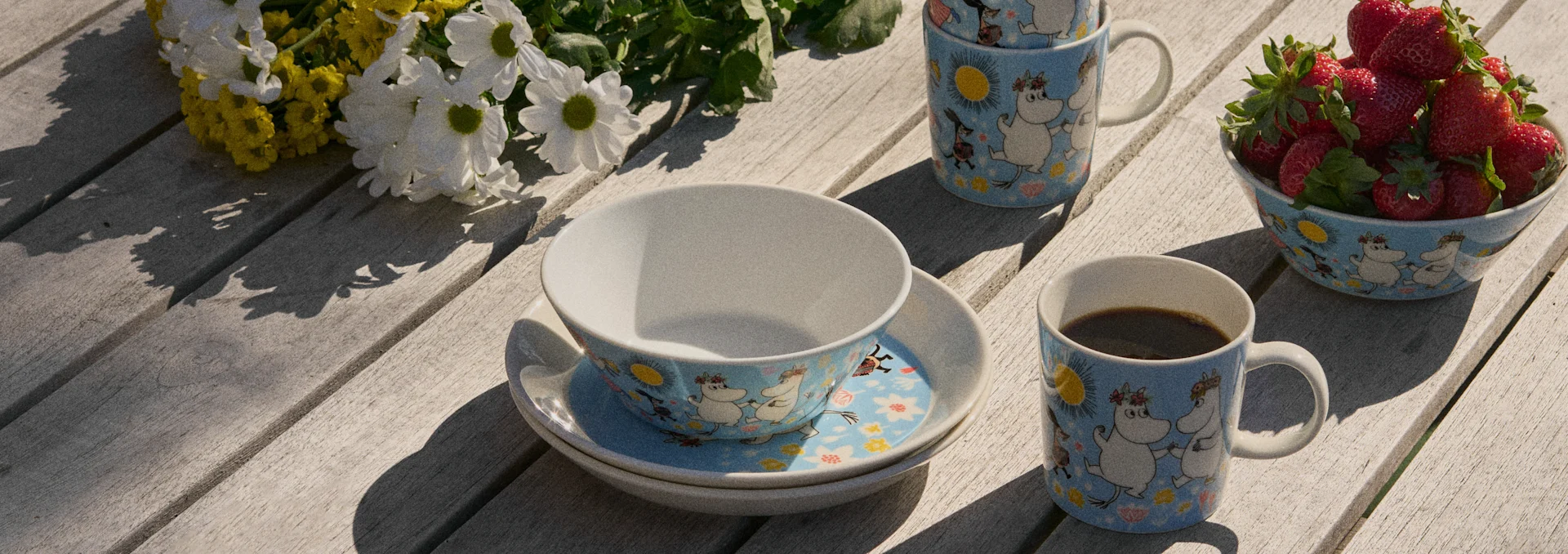 Muminmuggar, skålar och färska jordgubbar på ett soligt träbord bredvid vita och gula blommor.