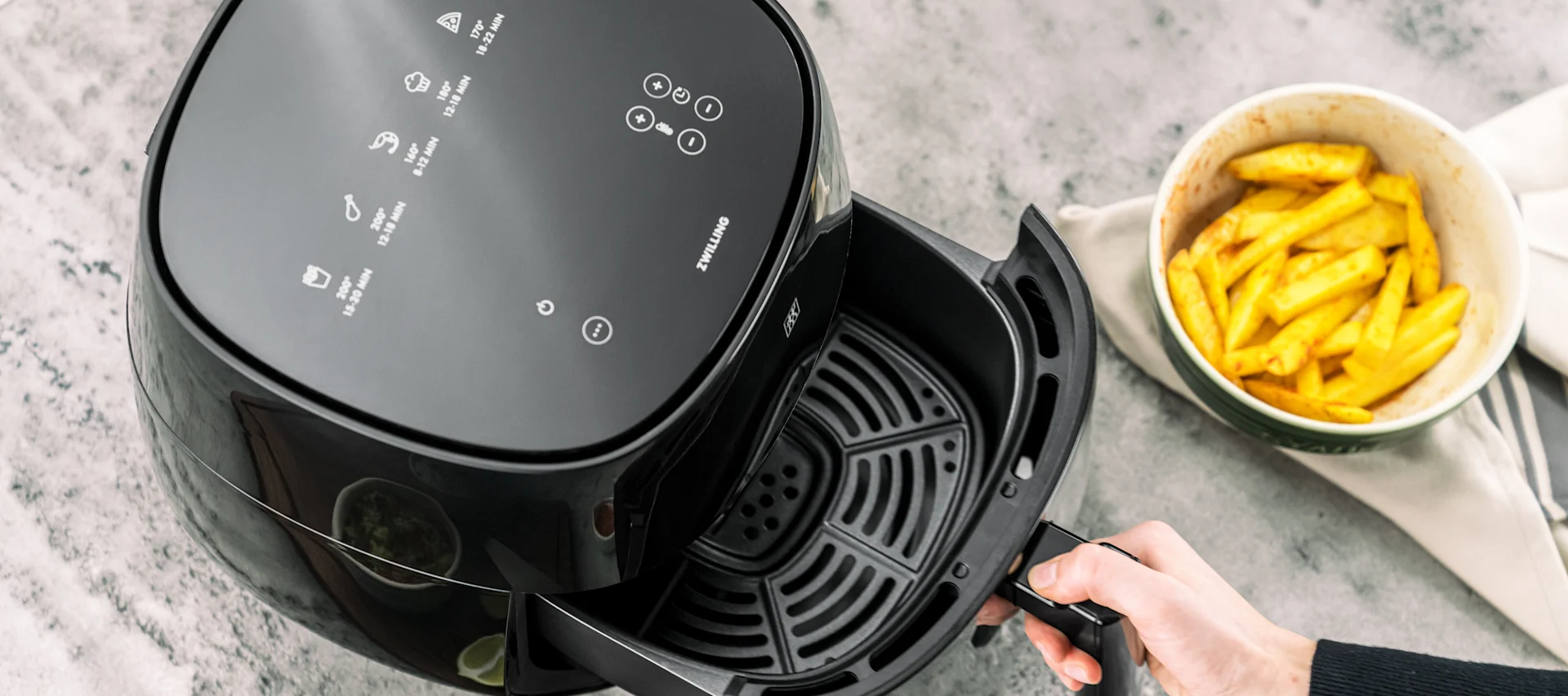 Fågelperspektiv av en hand som drar ut korgen från en svart airfryer, bredvid en skål med pommes frites.