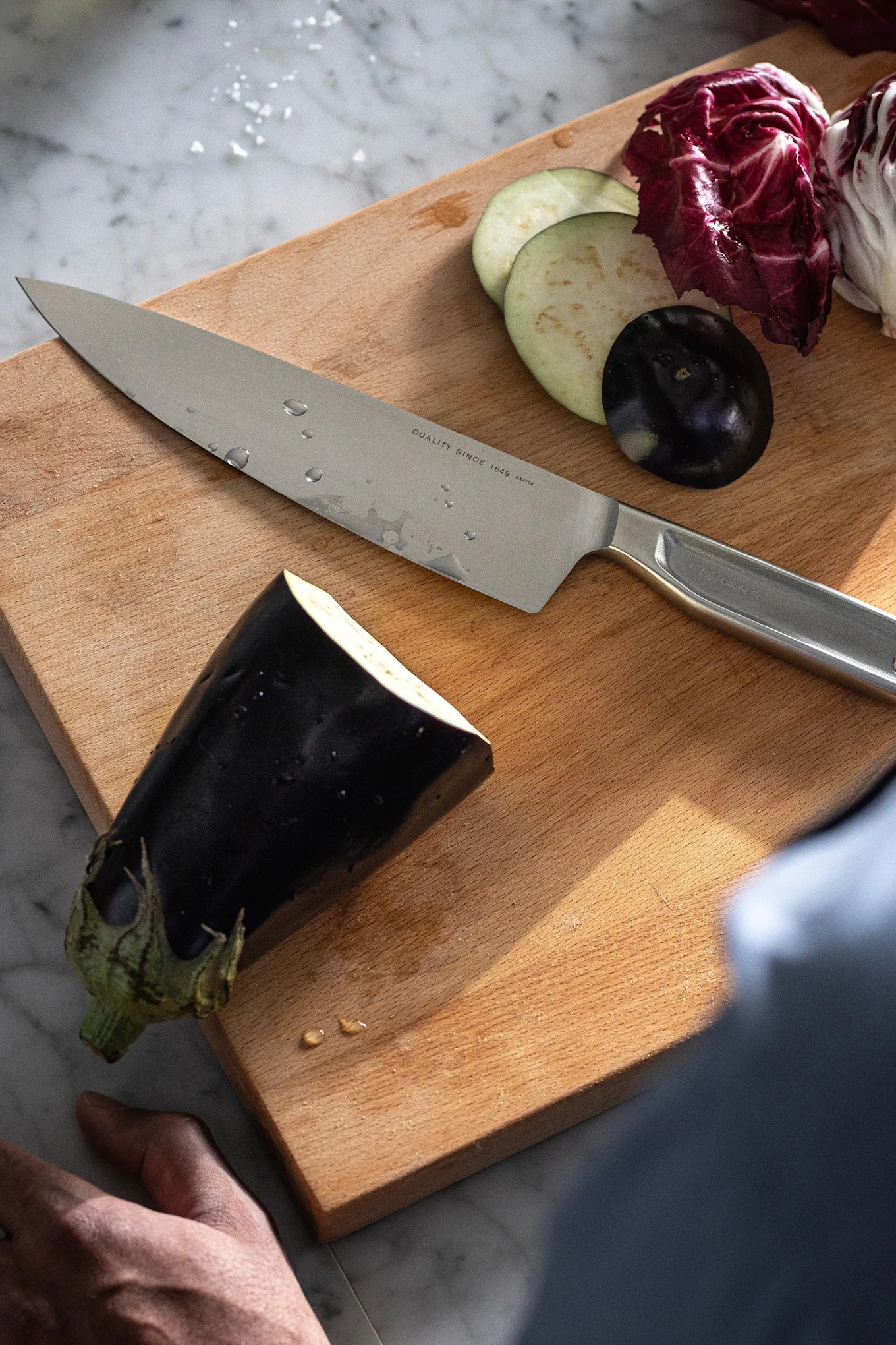 All Steel kökskniv från Fiskars ligger bredvid en aubergine på en skärbräda.
