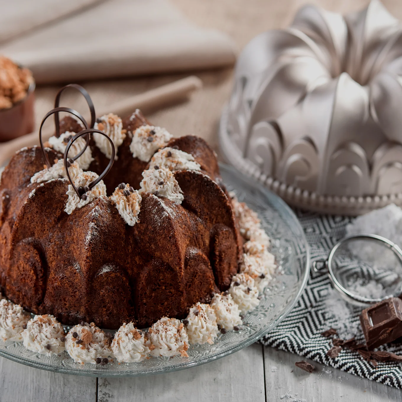 Choklad-bundtkaka med vispgrädde, chokladbitar och chokladlockar på ett glasfat, med en bundtform och ingredienser i bakgrunden.