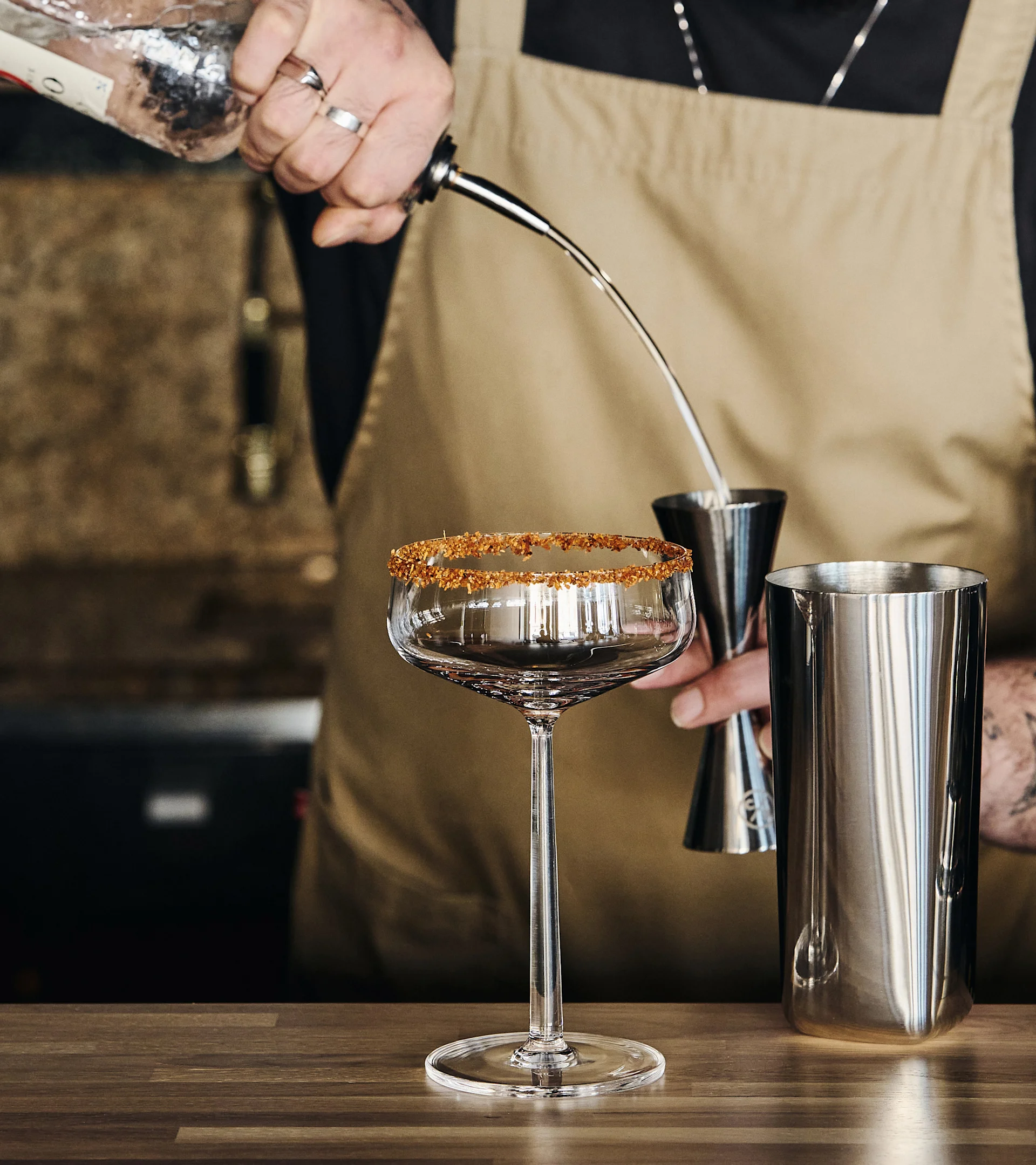 En bartender häller en drink från en flaska i ett mätglas, med ett sockerbelagt cocktailglas och en shaker på en träbänk.