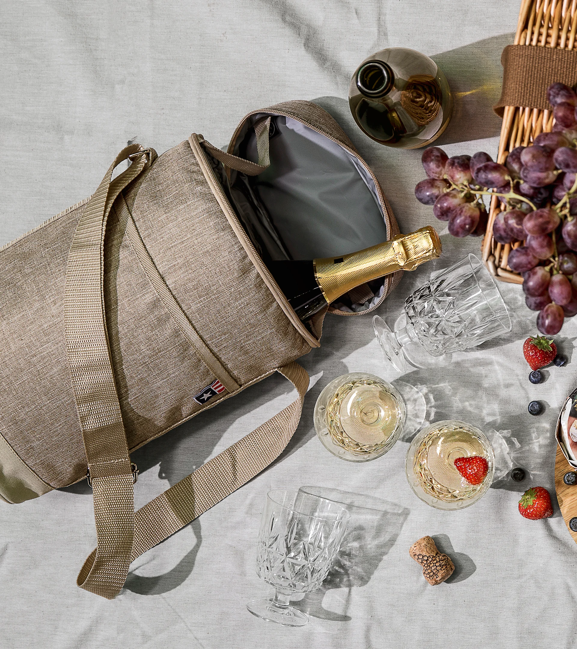Beige kylväska med champagne, mousserande vinglas, druvor och bär på en picknickfilt.