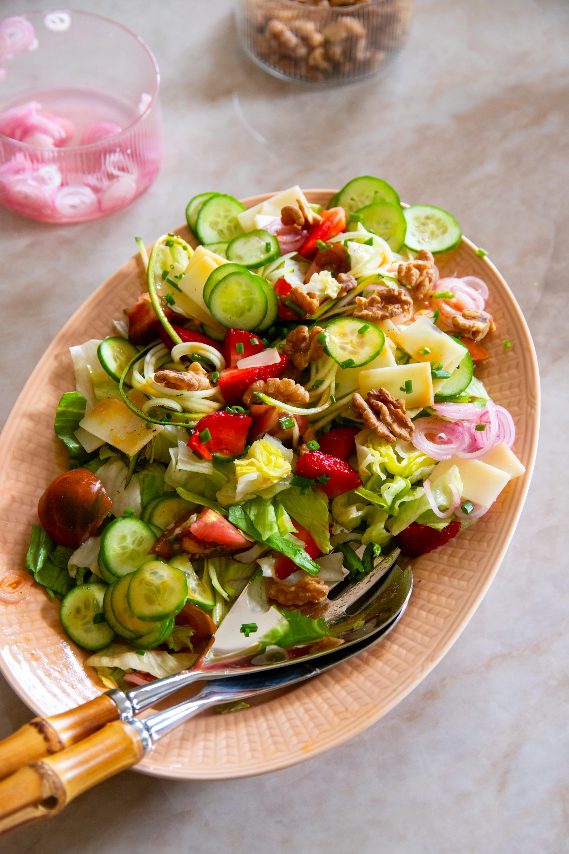 Färgrik sallad på ovalt fat: sallad, gurka, jordgubbar, ost, valnötter, rödlök, med serveringsbestick av bambu.