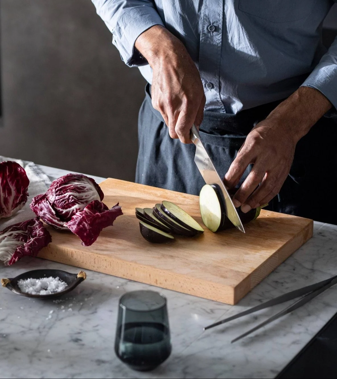 Närbild av en person som skivar aubergine på en träskärbräda med radicchio och salt i närheten.