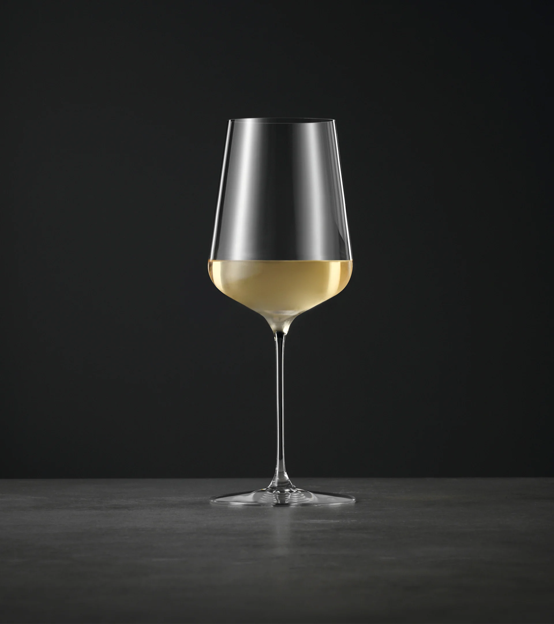 Ett högt, elegant vinglas med lång stjälk, halvfyllt med ljust vitt vin, mot en mörk bakgrund.