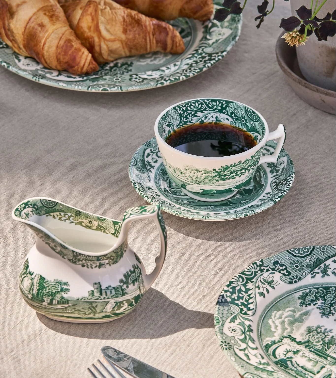Frukostbord med grön-vit mönstrad porslinskopp med kaffe, gräddkanna och tallrikar med croissanter.