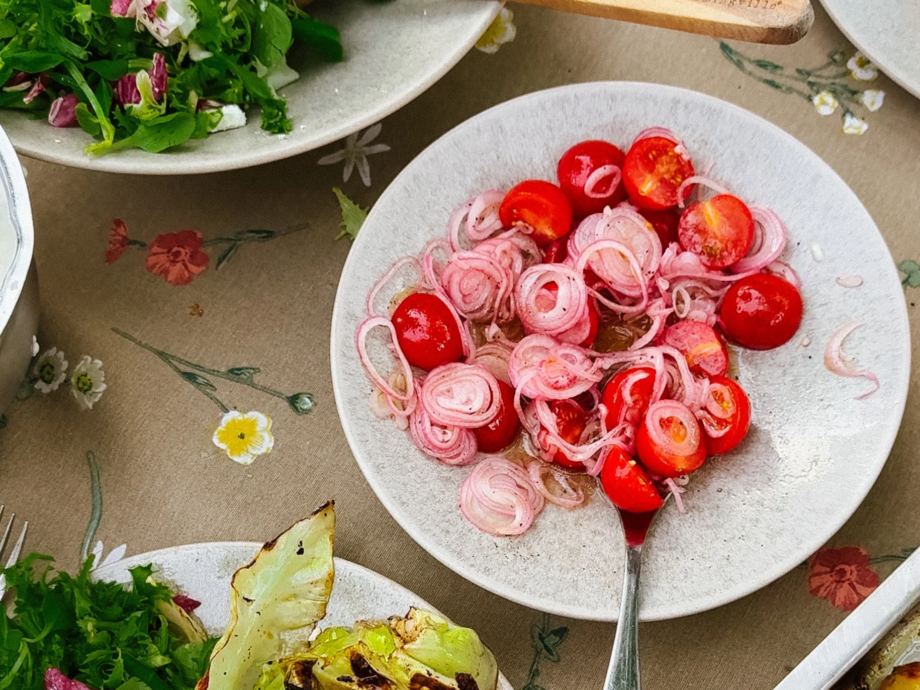 Närbild av en tallrik med körsbärstomat- och rödlökssallad, flankerad av grönsallad och grillad kål på en blommig duk.