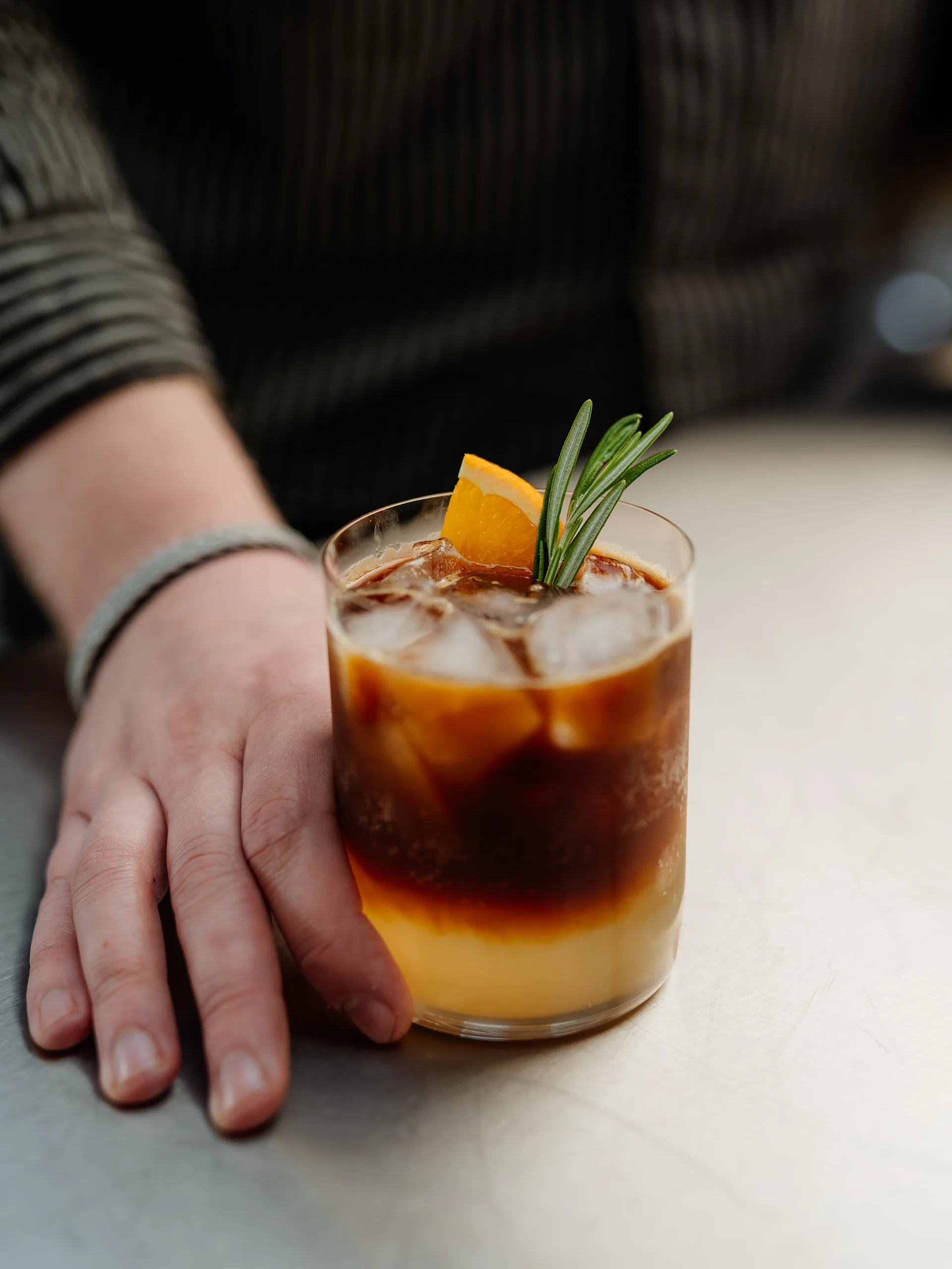 Hand håller en iskaffe- och apelsinjuicecocktail med rosmarin och en apelsinskiva som garnering.