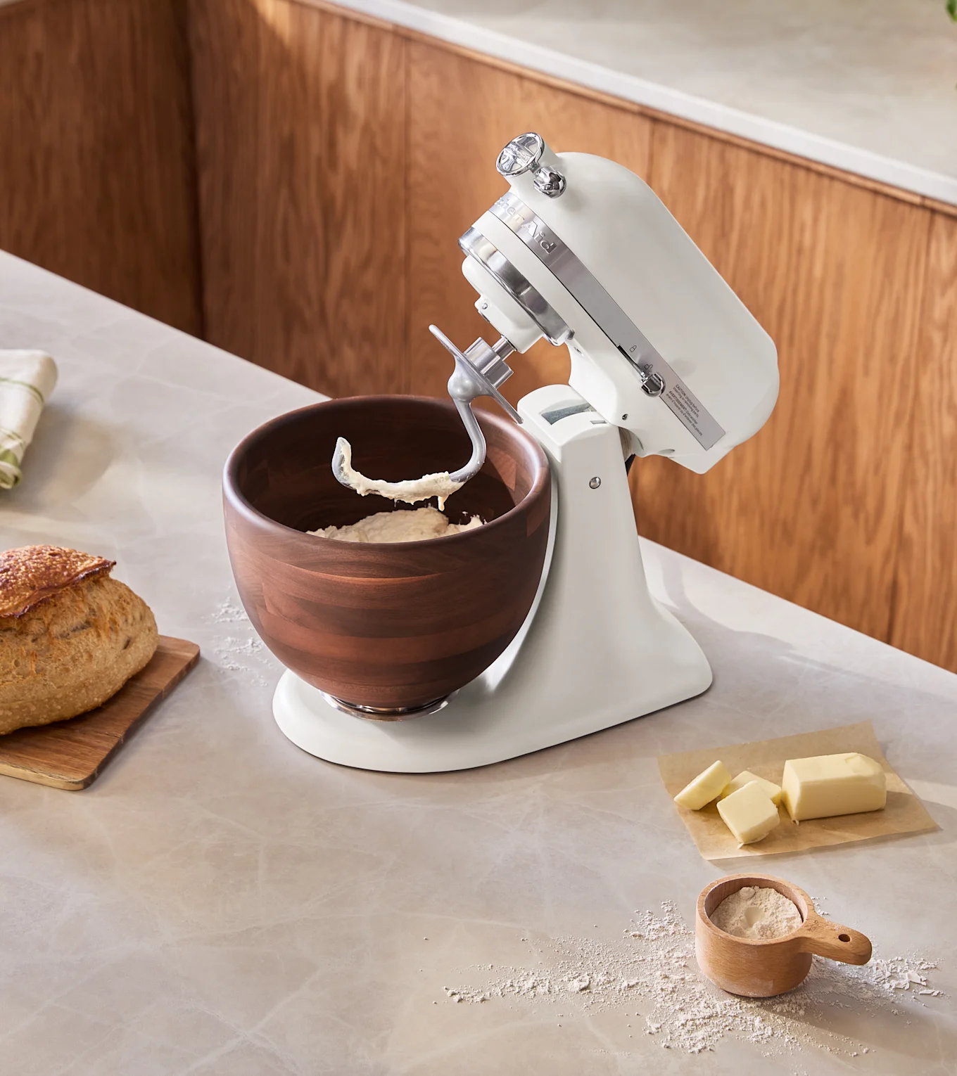 En vit köksmaskin med en mörk träskål med deg på en köksbänk. Ett färskt bröd, mjöl i en kopp och smör ligger i närheten.