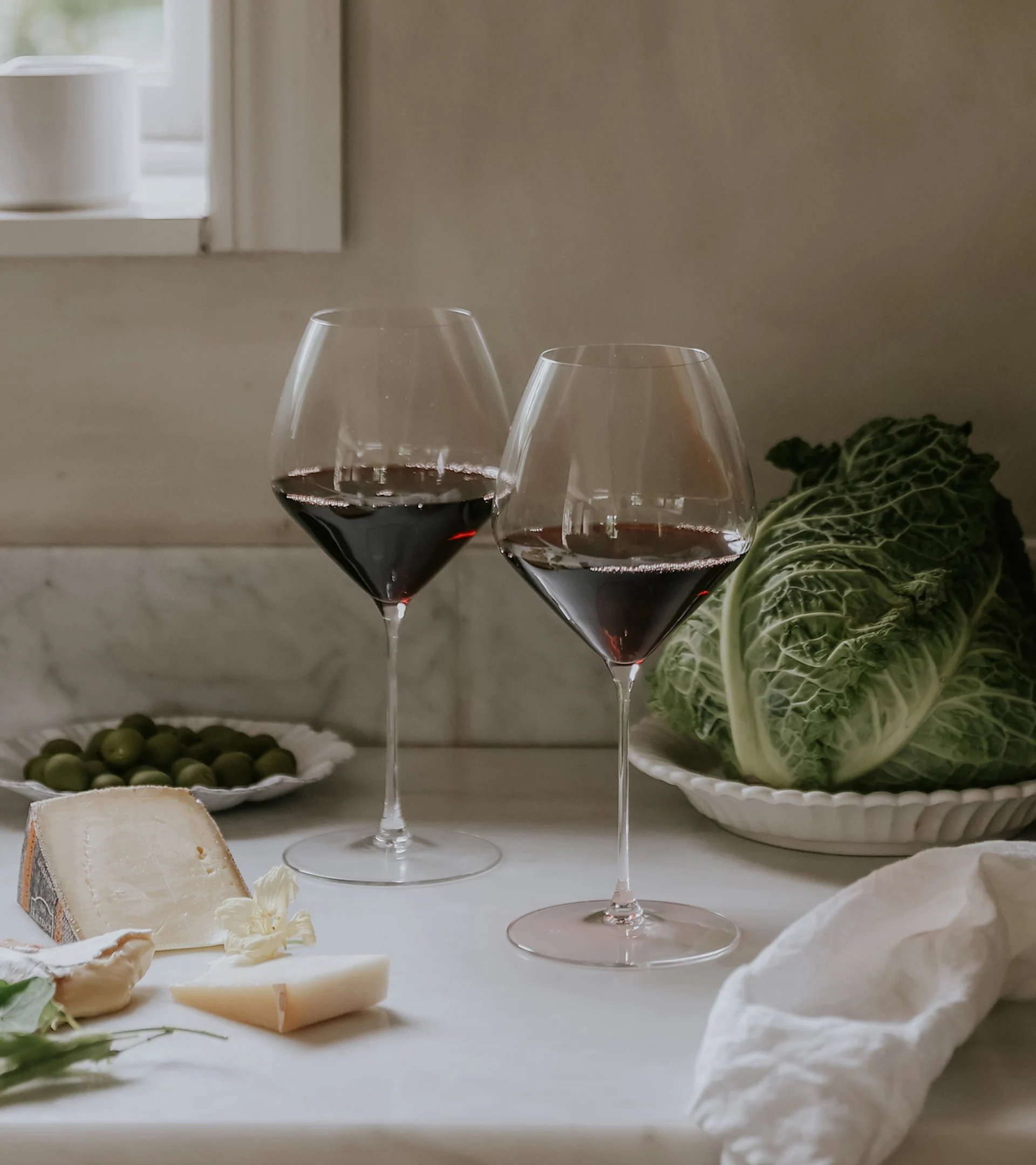 Två glas rött vin, diverse ostar, oliver och ett savojkålhuvud på en köksbänk i marmor.