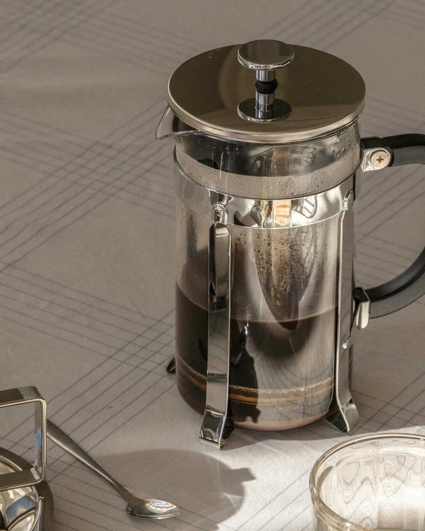 French press kaffebryggare med mörkt kaffe på en randig duk, bredvid en sked och ett glas.