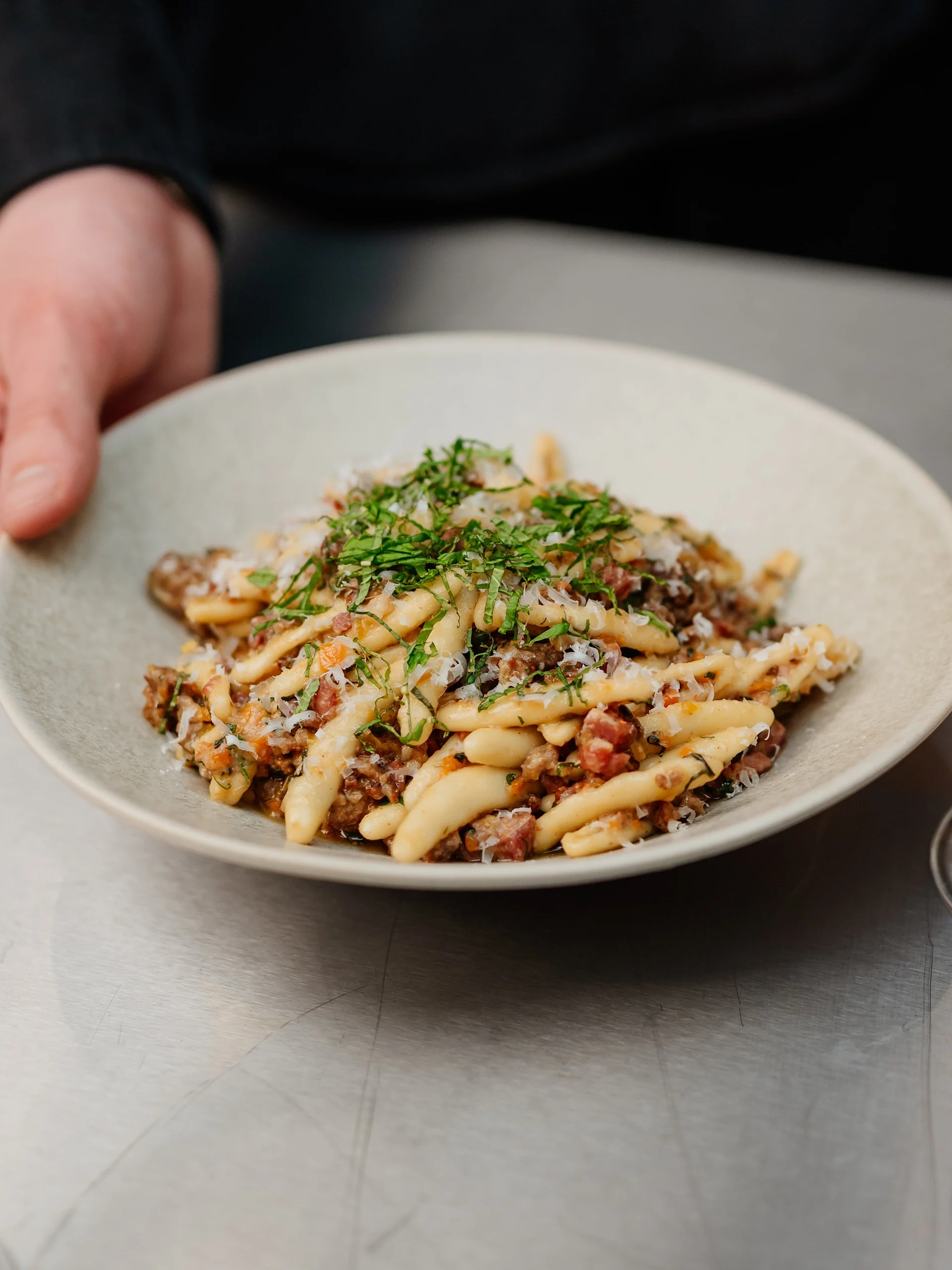 En hand håller en tallrik med vriden pasta i köttfärssås, riven ost och färska örter.