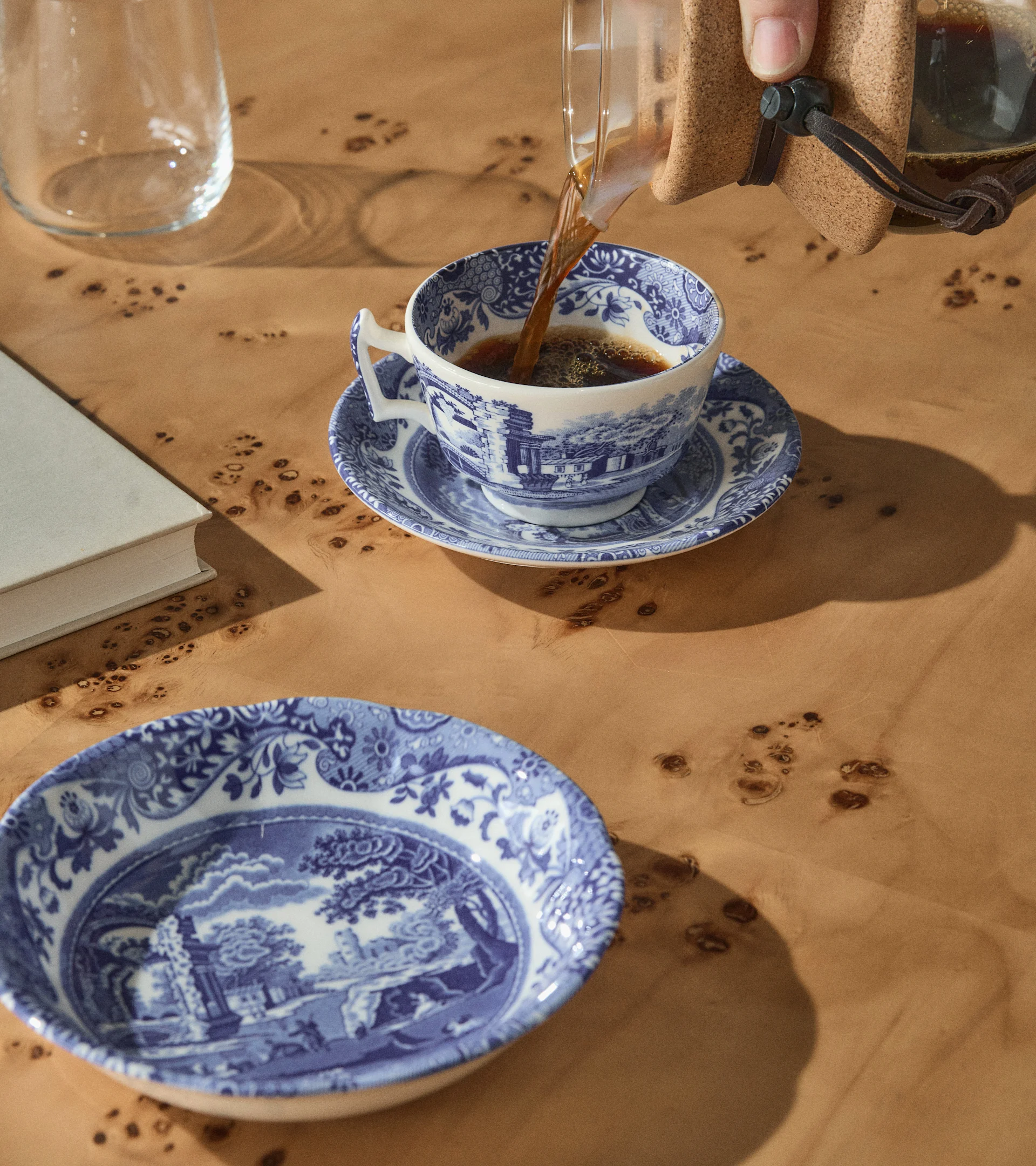 En hand häller kaffe i en blå och vit mönstrad tekopp på ett träbord med en matchande skål, en bok och ett glas.