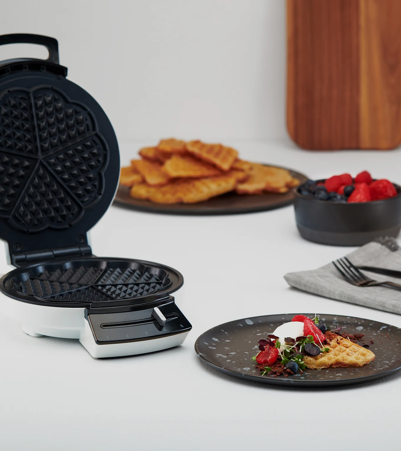 Öppet våffeljärn bredvid tallrik med våffla, grädde, bär och mikrogrönt. Fler våfflor och bär i bakgrunden.