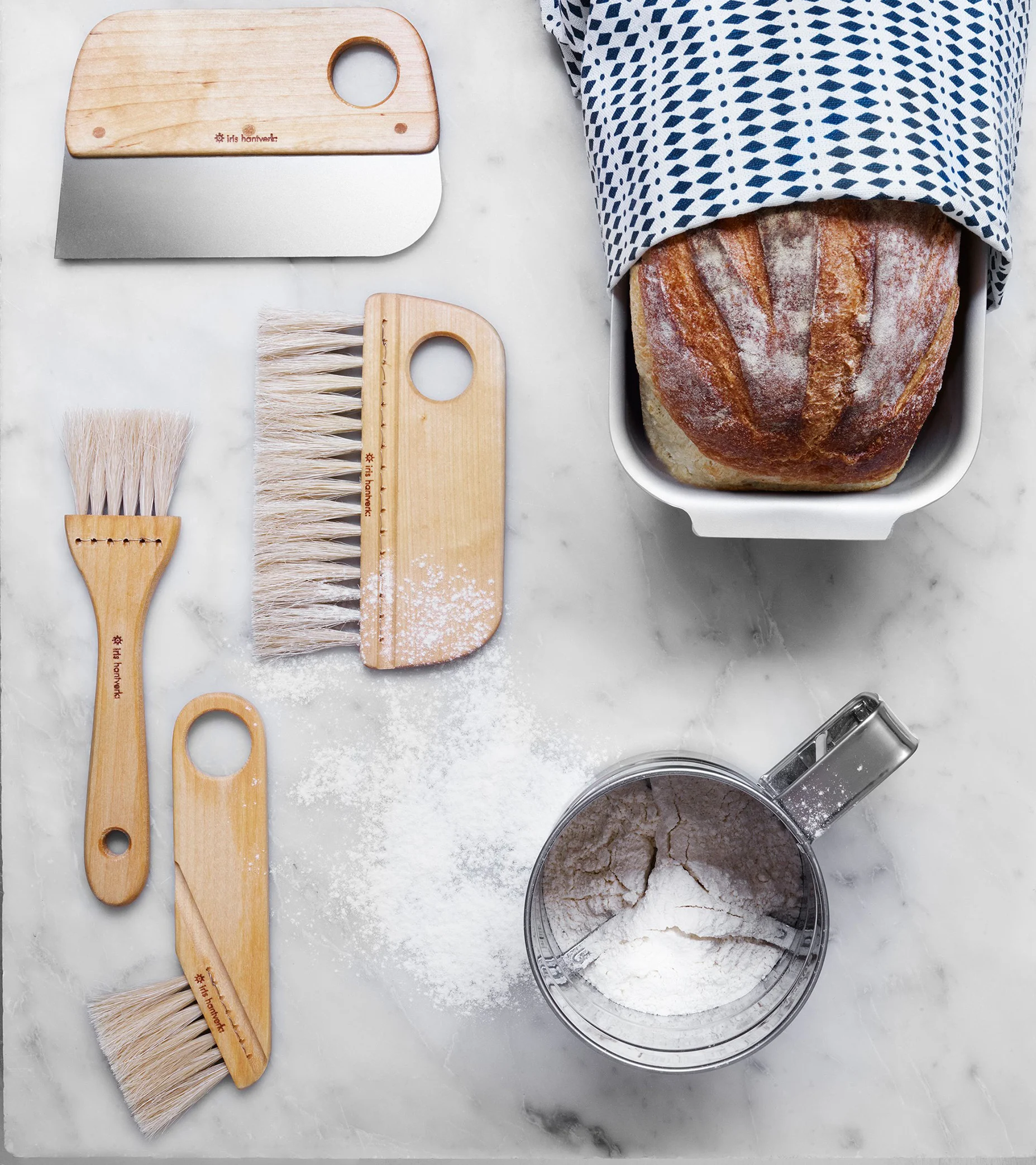 Bakredskap och ett nybakt bröd på en marmorbänkskiva. Inkluderar degskrapa, borstar, mjölsikt och spritt mjöl.