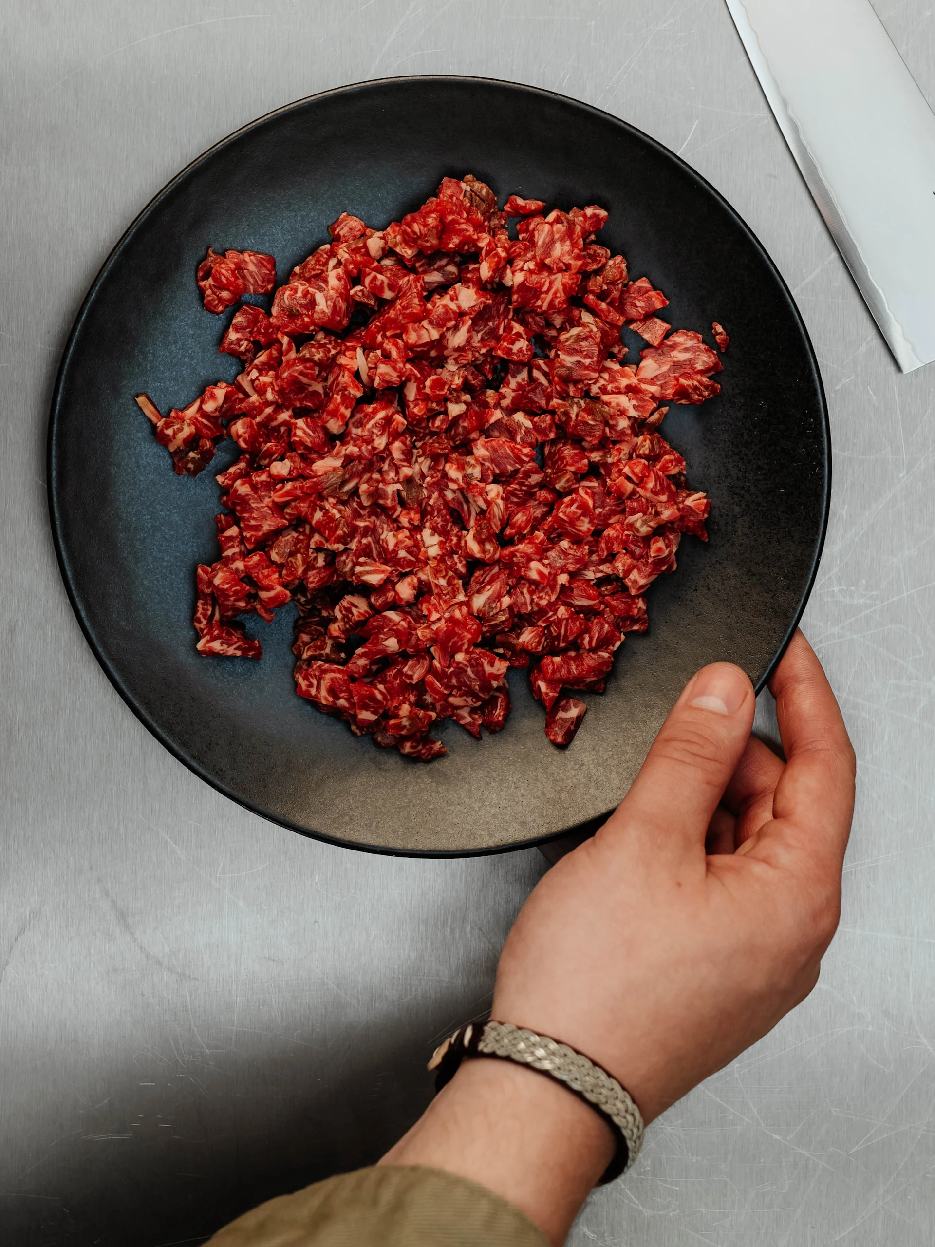 En hand håller en svart tallrik med tärnat rått rött kött på en repig metallyta, med ett knivblad.