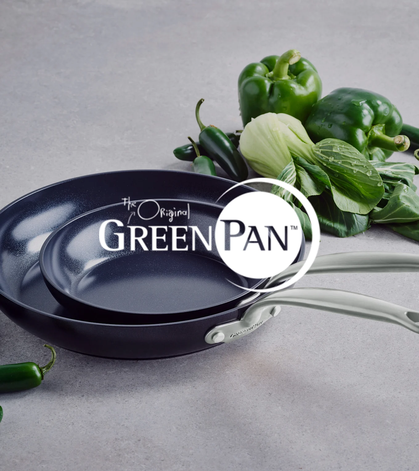 Två mörka non-stick pannor med ljusa handtag, staplade, bredvid grön paprika, pak choi och jalapeños. GreenPan logotyp.