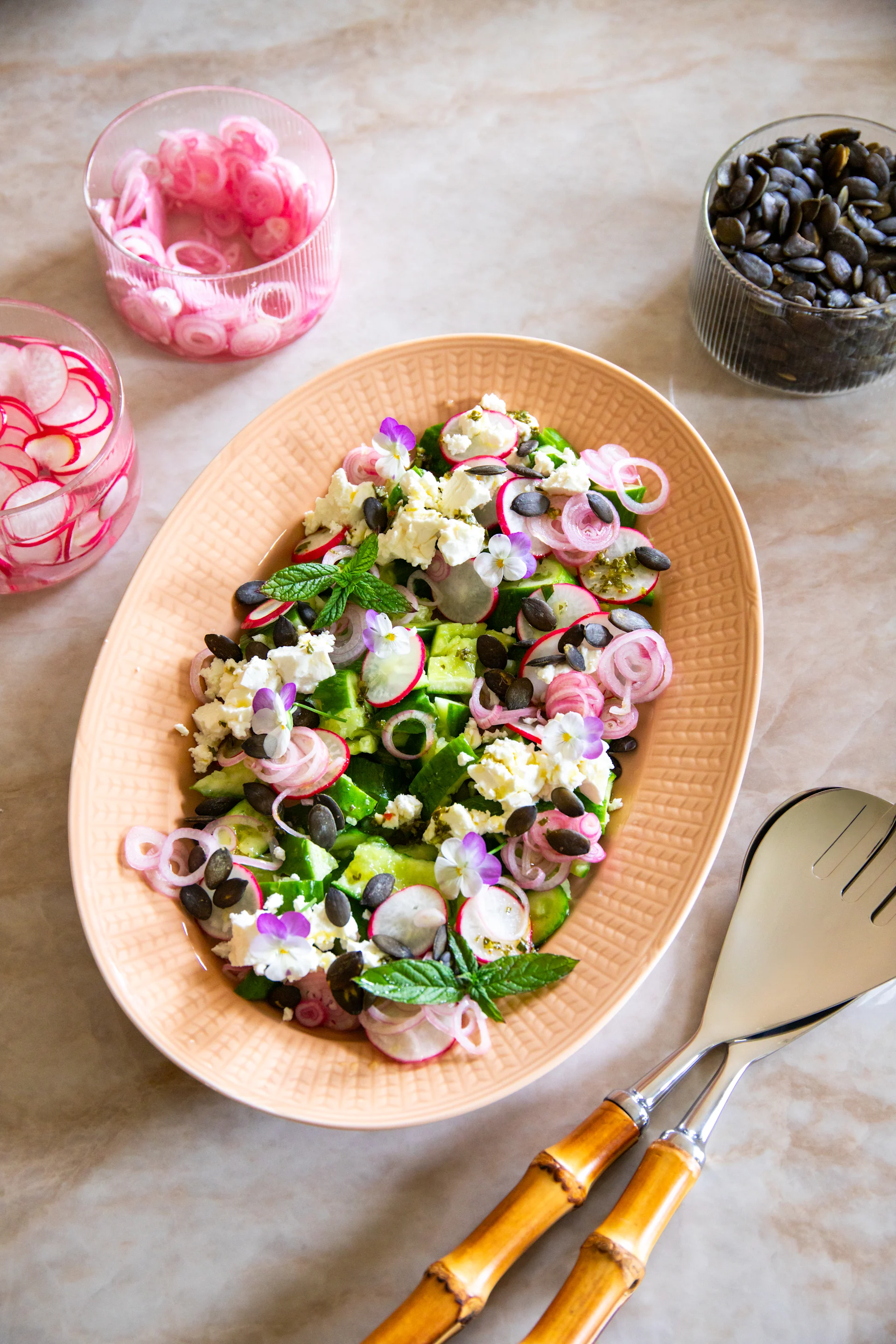 Färgrik sallad på persikofärgad tallrik: gurka, rädisa, lök, feta, pumpafrön, mynta, blommor. Bestick.