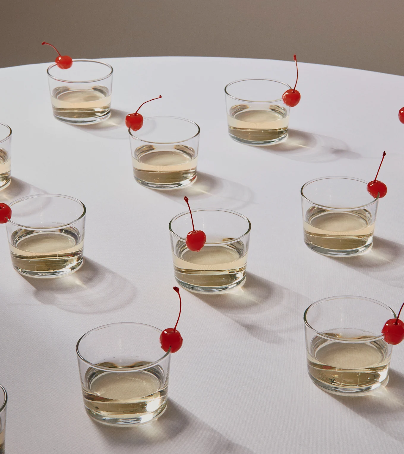 Flera glas med ljusa drycker, var och en med en röd körsbärsgarnityr, på en vit yta.