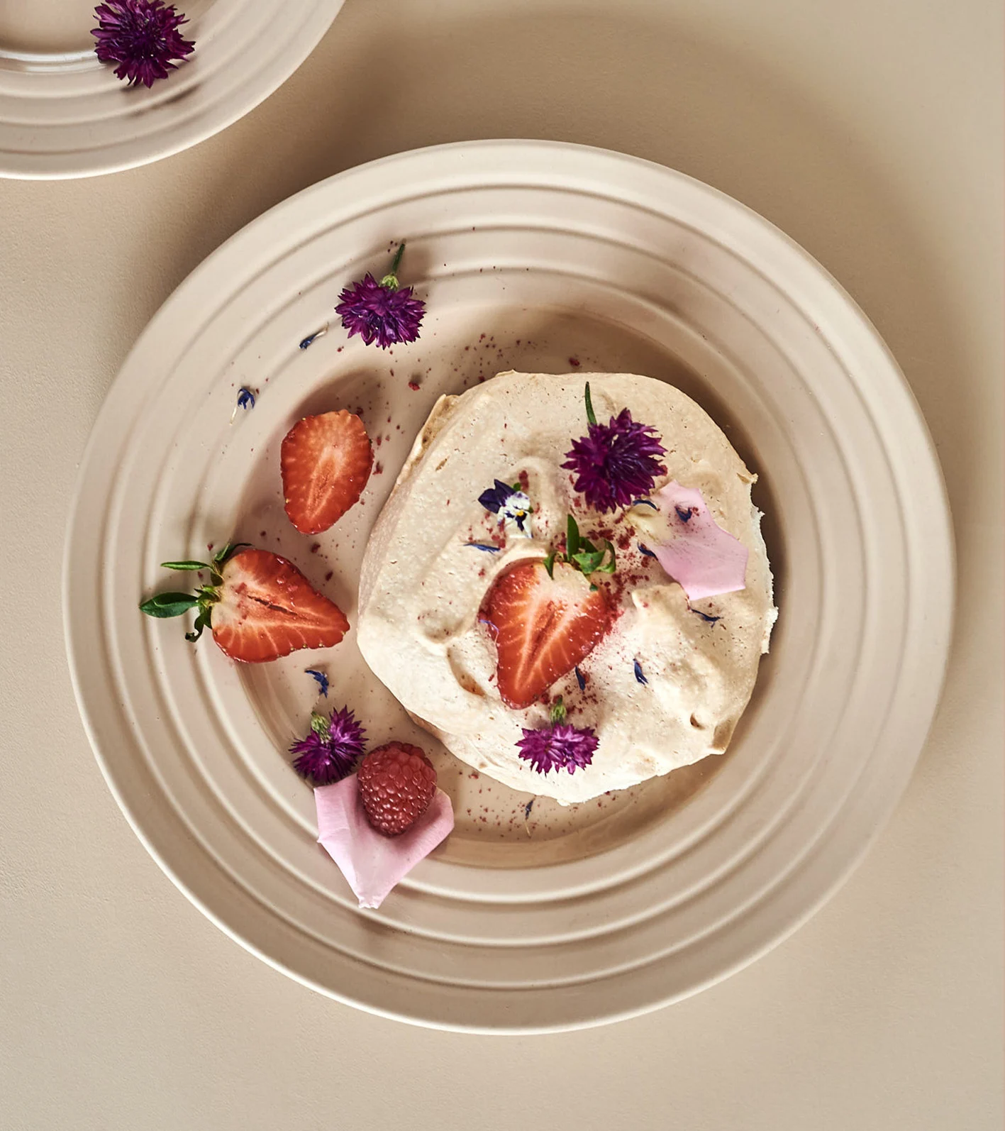 En krämig dessert med jordgubbar, ett hallon och lila blommor på en beige räfflad tallrik.