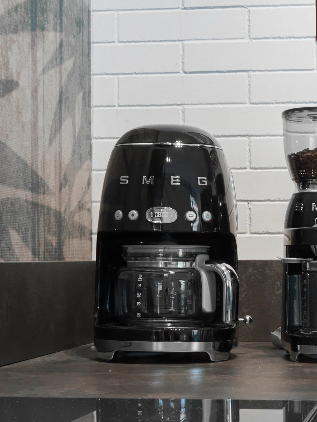 Svart Smeg kaffebryggare med glaskanna och digital display som visar '08:00' på en mörk bänkskiva
