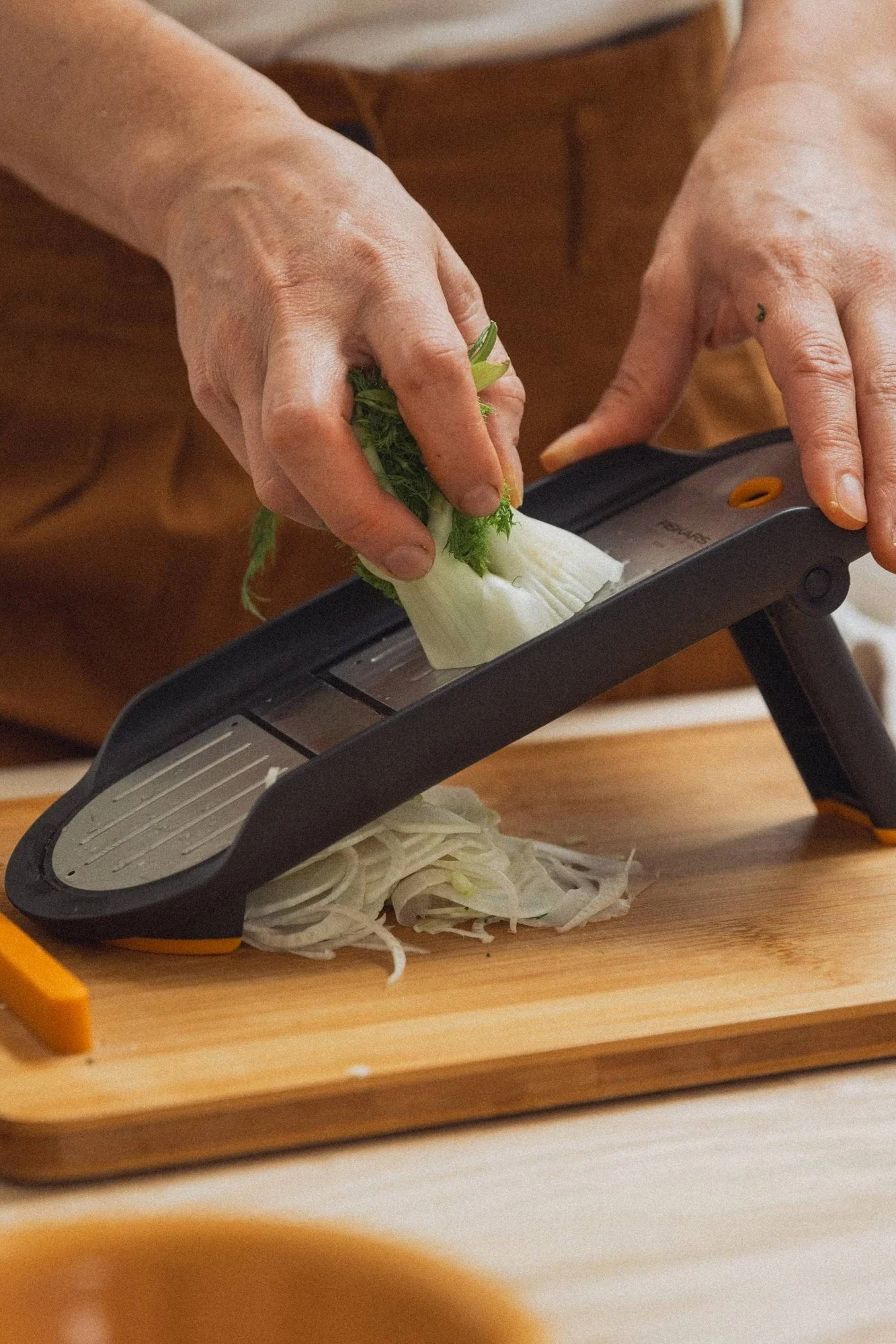 Händer skivar fänkål med en grå mandolin på en skärbräda i trä.