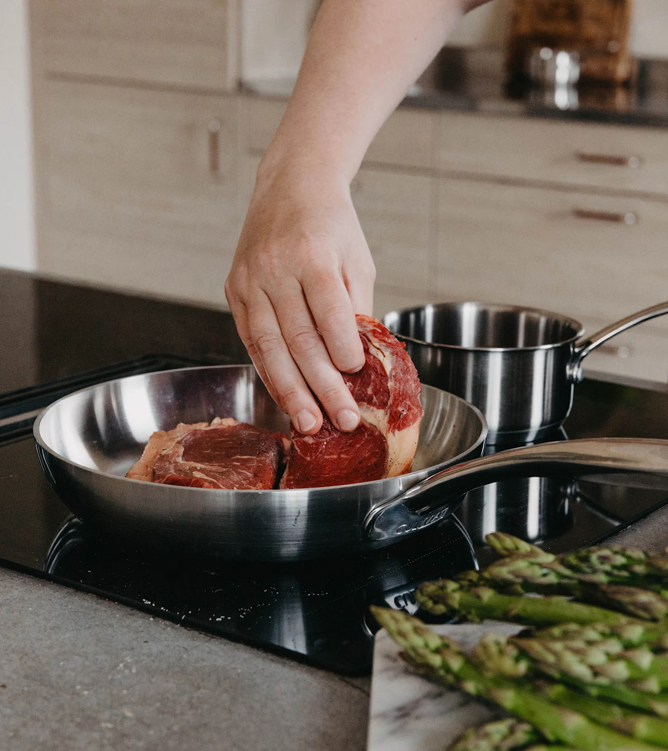 Hand lägger rå biff i stekpanna av rostfritt stål på häll, med sparris på marmorbräda i förgrunden.