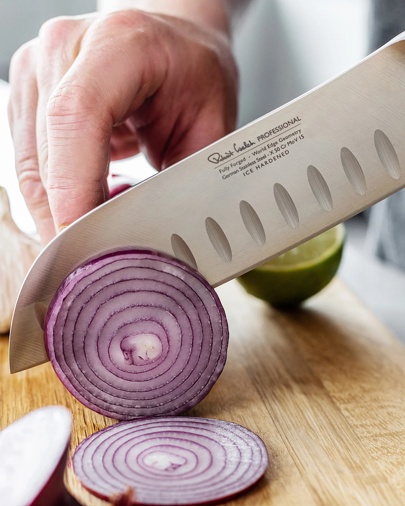 En kockkniv med texturerat blad skivar en rödlök på en träskärbräda, med en lime i bakgrunden.