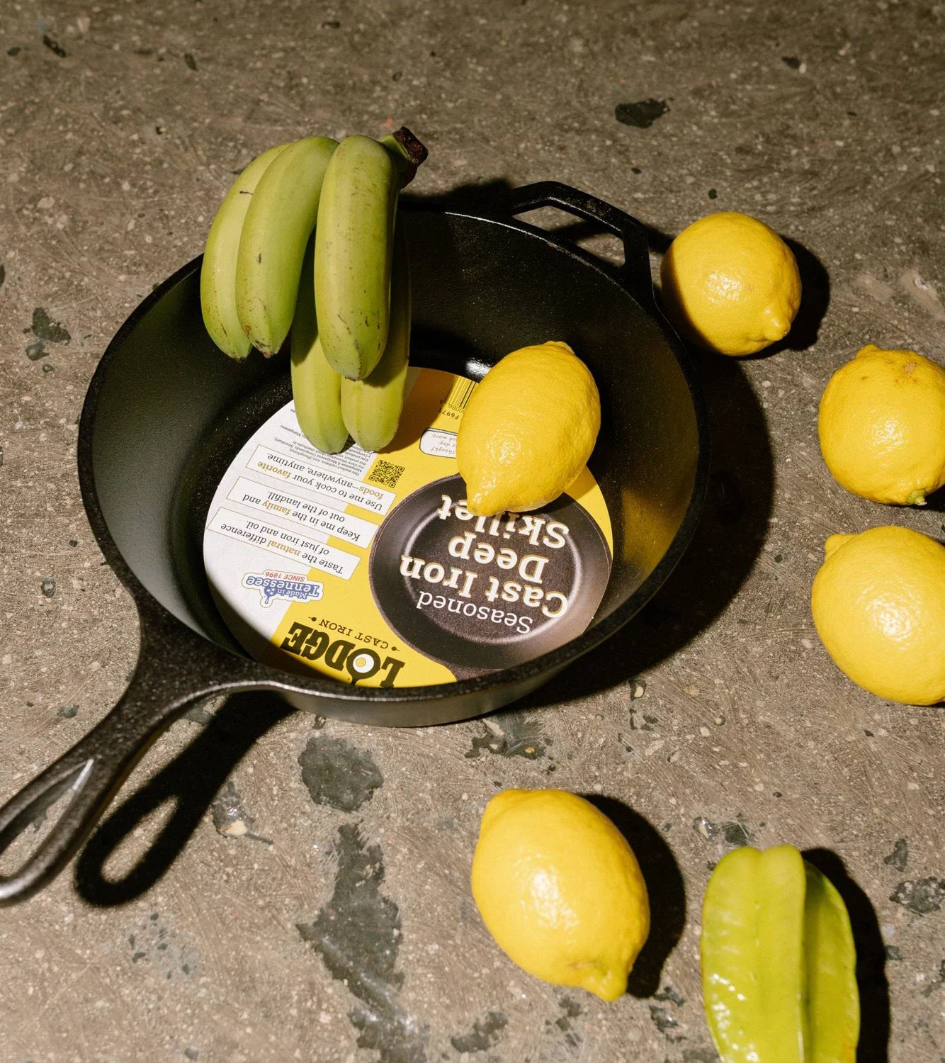 Gjutjärnspanna med etikett inuti, omogna bananer, citroner och en stjärnfrukt på en mörk yta.