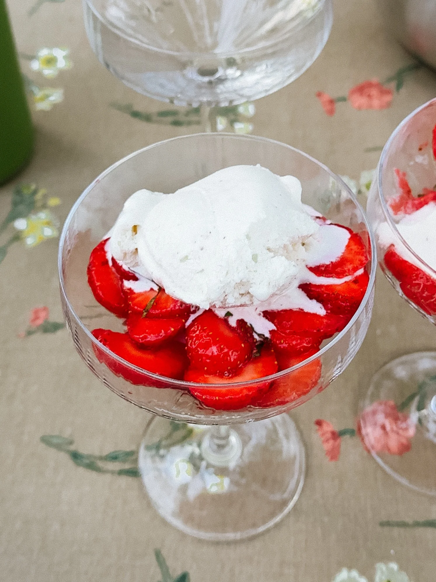 En glasdessertskål fylld med skivade röda jordgubbar och en skopa vit glass, på en blommig duk.
