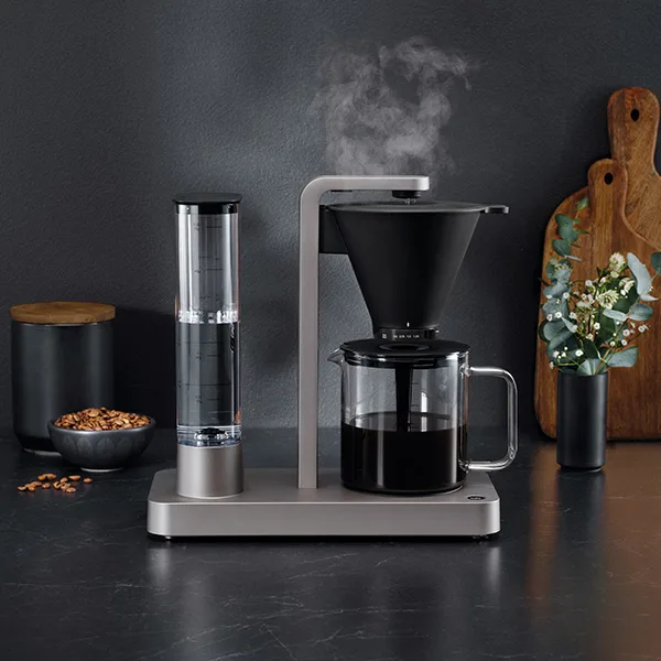 Kaffebryggare med kaffekanna, två kaffekoppar, skål med kaffebönor och kakor på mörk bänkskiva.