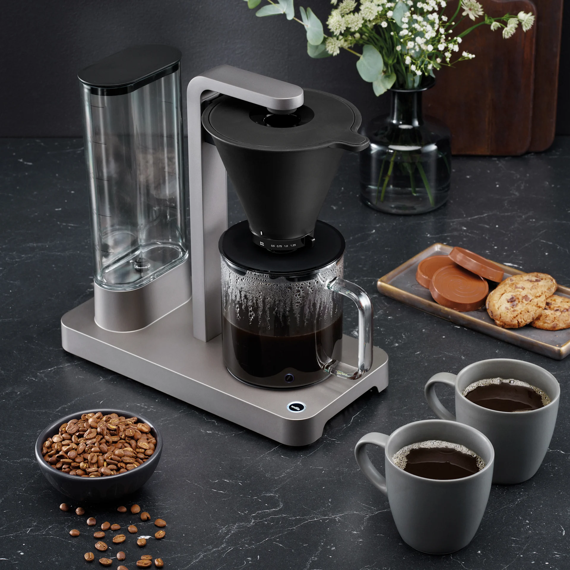 Kaffebryggare med kaffekanna, två kaffekoppar, skål med kaffebönor och kakor på mörk bänkskiva.