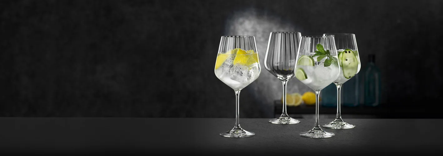 Fyra stjälkade glas på mörk bänk, två med garnerade drinkar och is, två tomma.