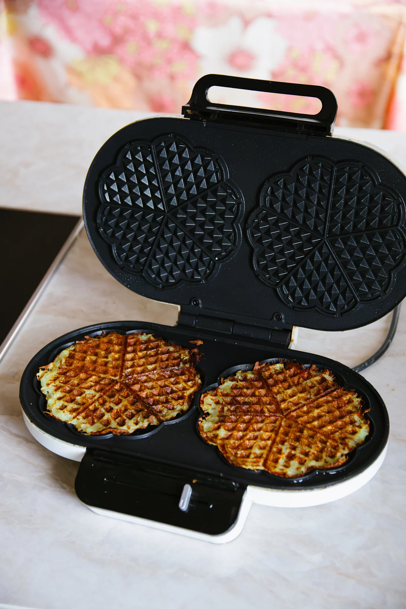 Två gyllenbruna, hjärtformade våfflor som tillagas i ett öppet vitt och svart våffeljärn.