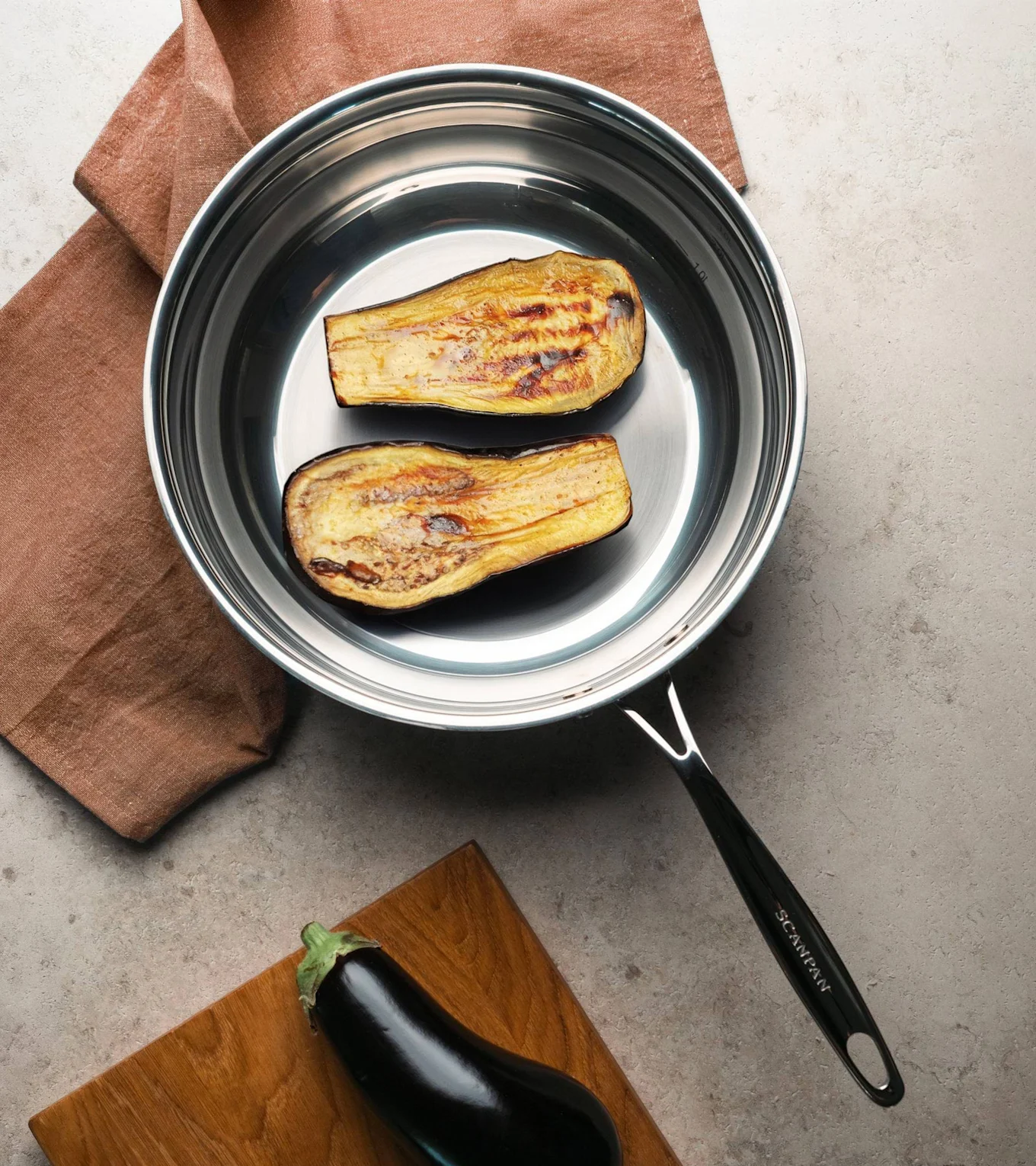 Två grillade auberginehalvor i en stekpanna av rostfritt stål, med en rå aubergine på en skärbräda intill.