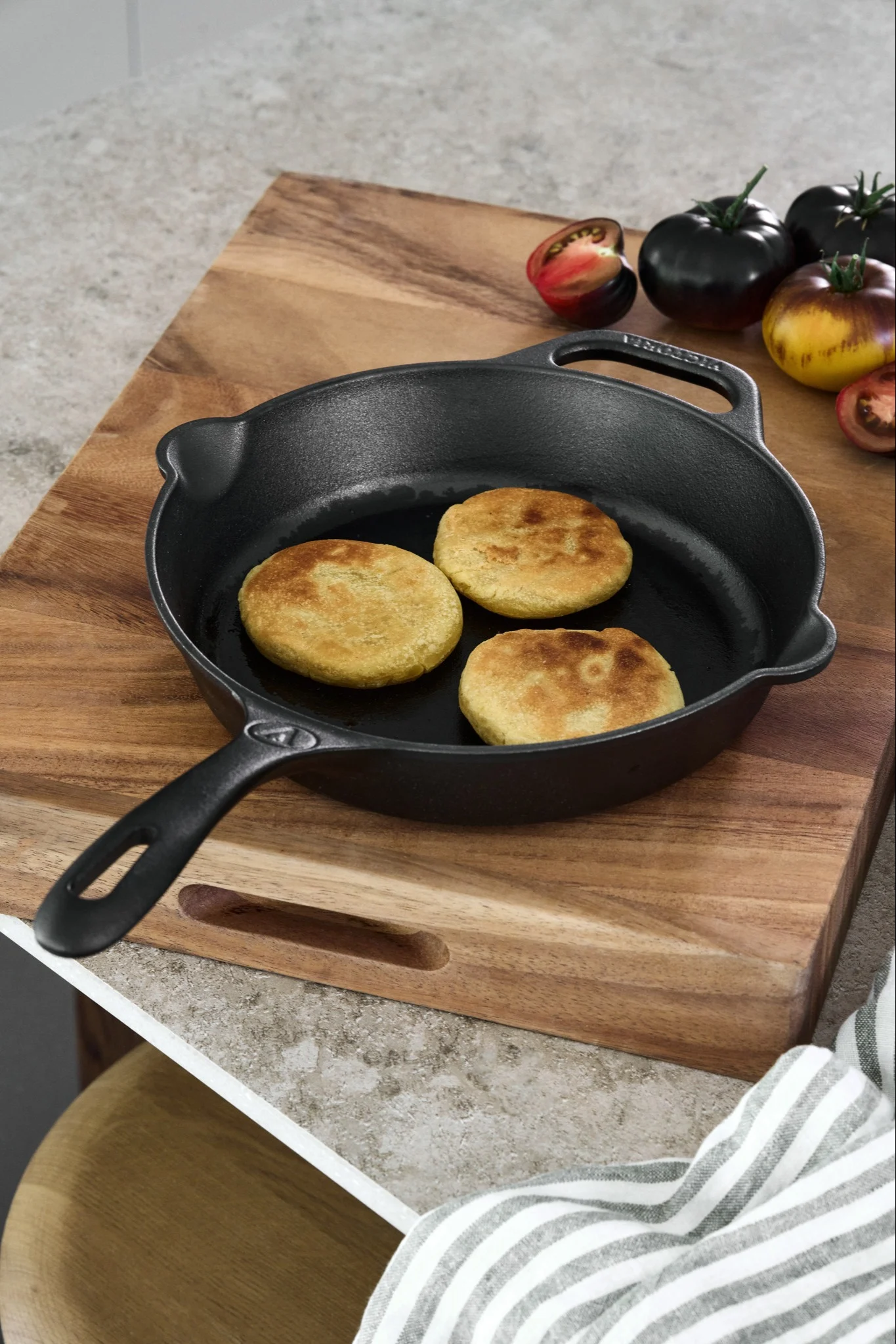 En gjutjärnspanna placeras på en träskärbräda med en samling tomater och stekta arepas. 