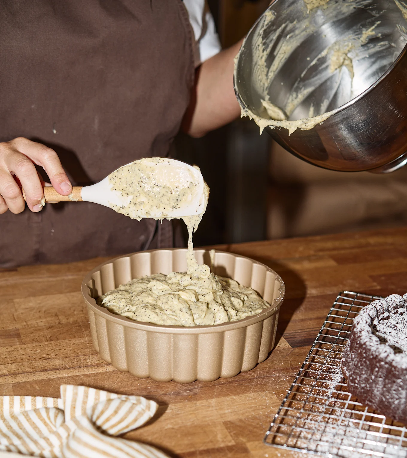 Person skopar vallmofrödeg i en guldfärgad räfflad bakform. En chokladkaka med florsocker finns i bakgrunden.