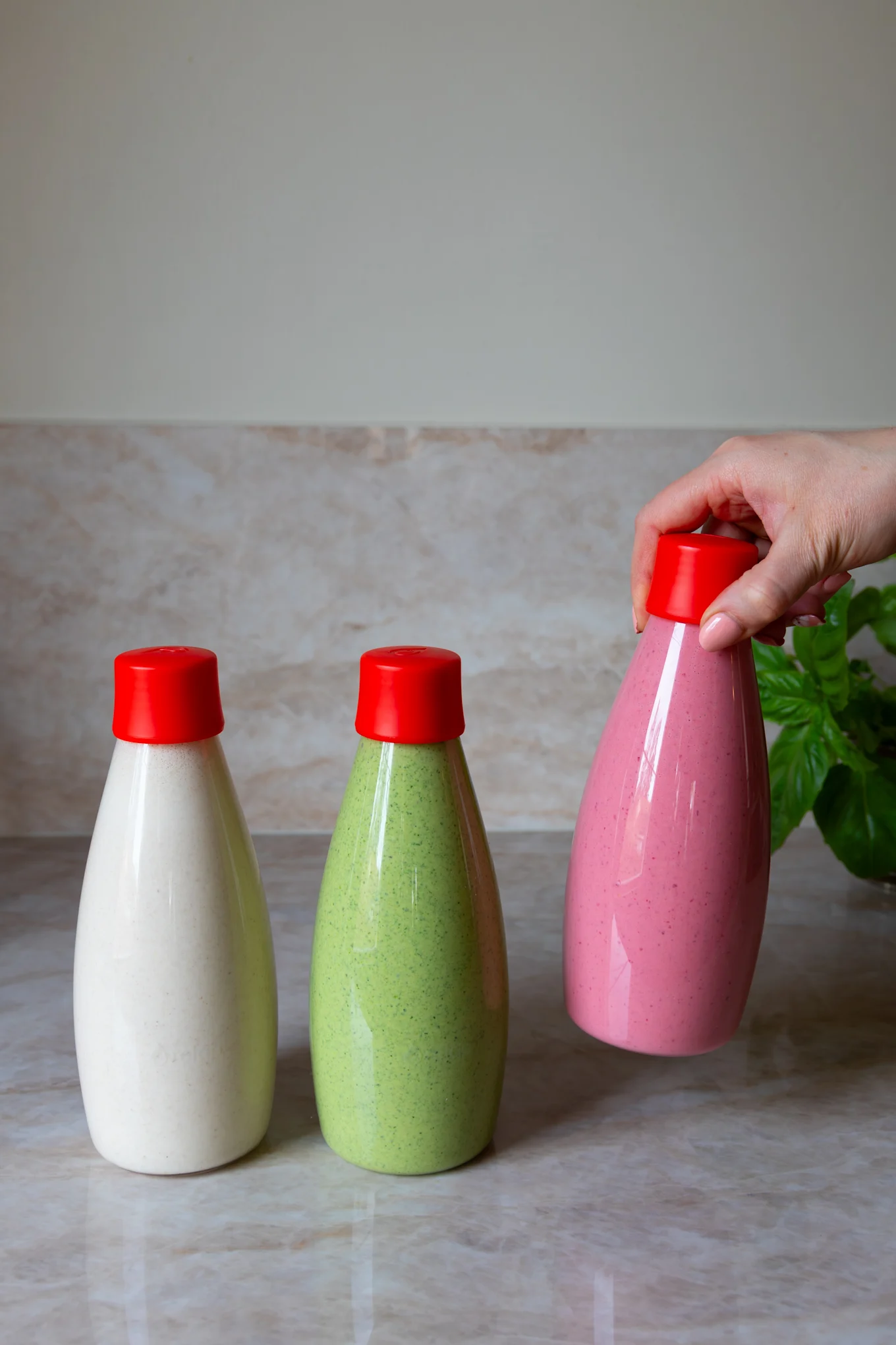 Tre flaskor med röda korkar, innehållande beige, grön och rosa vätska, där en hand håller den rosa flaskan.