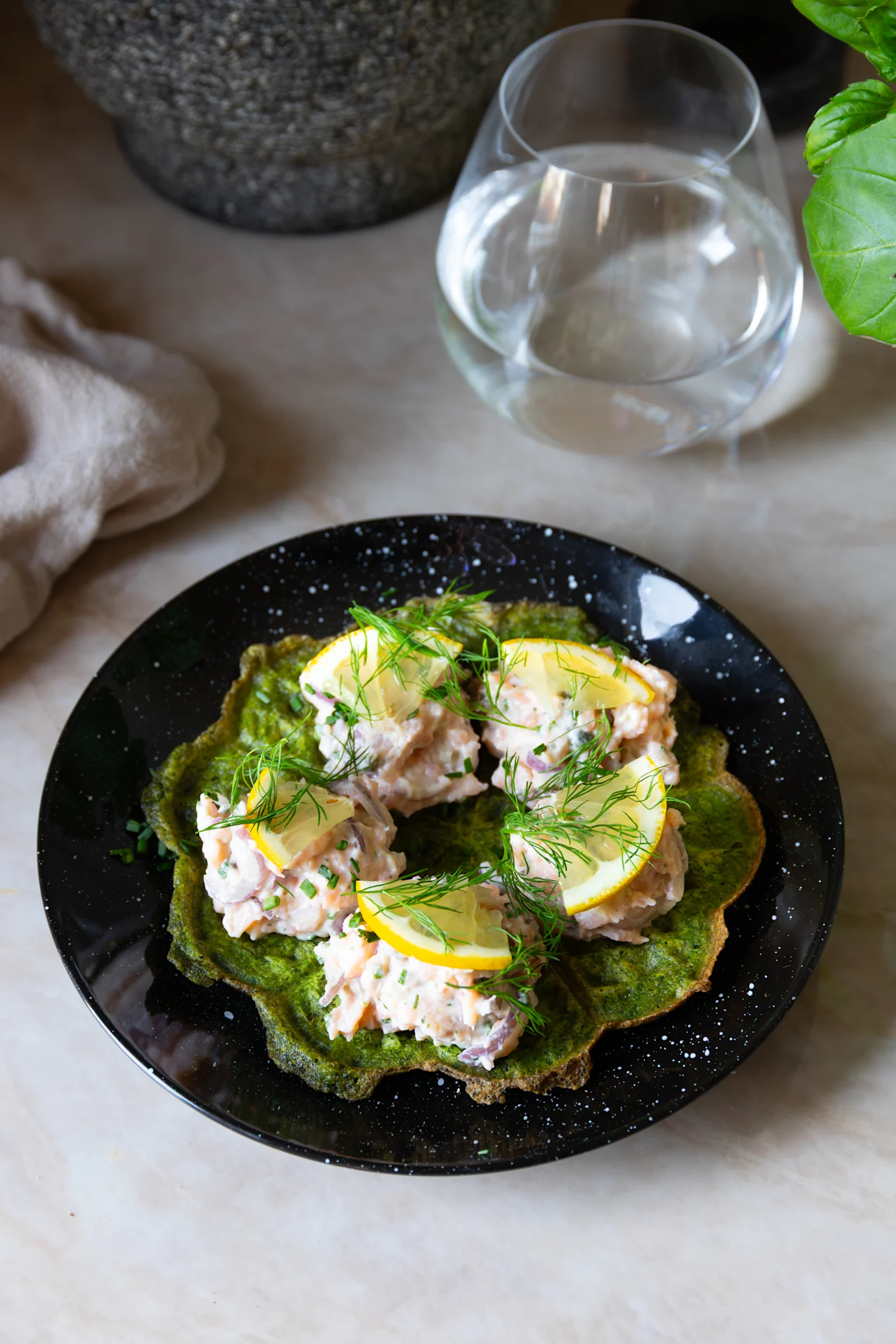 Grön våffla med laxröra, citronskivor och färsk dill på en svart, fläckig tallrik.