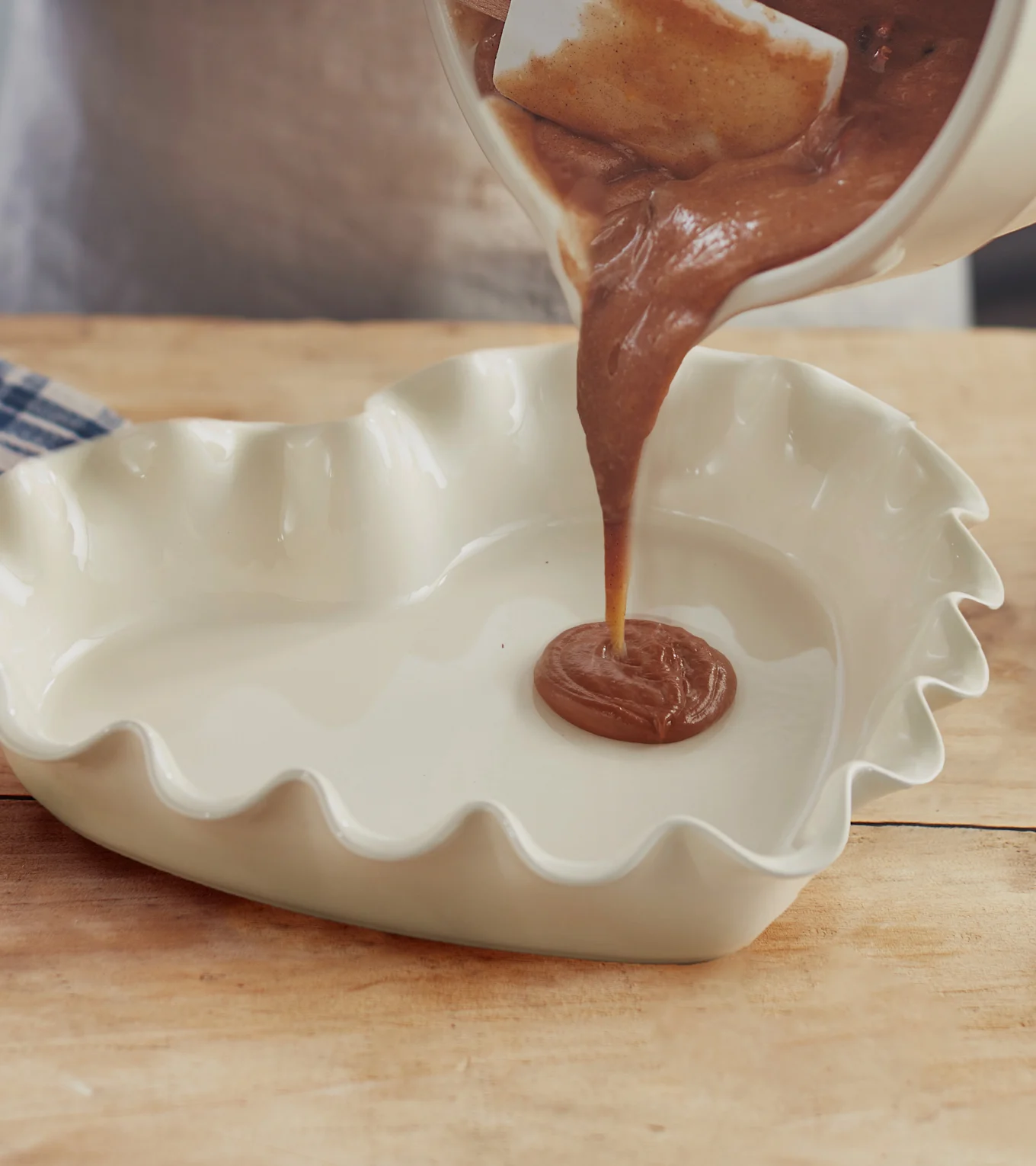 Häller chokladsmet från en skål i en vit hjärtformad ugnsform på ett träbord.