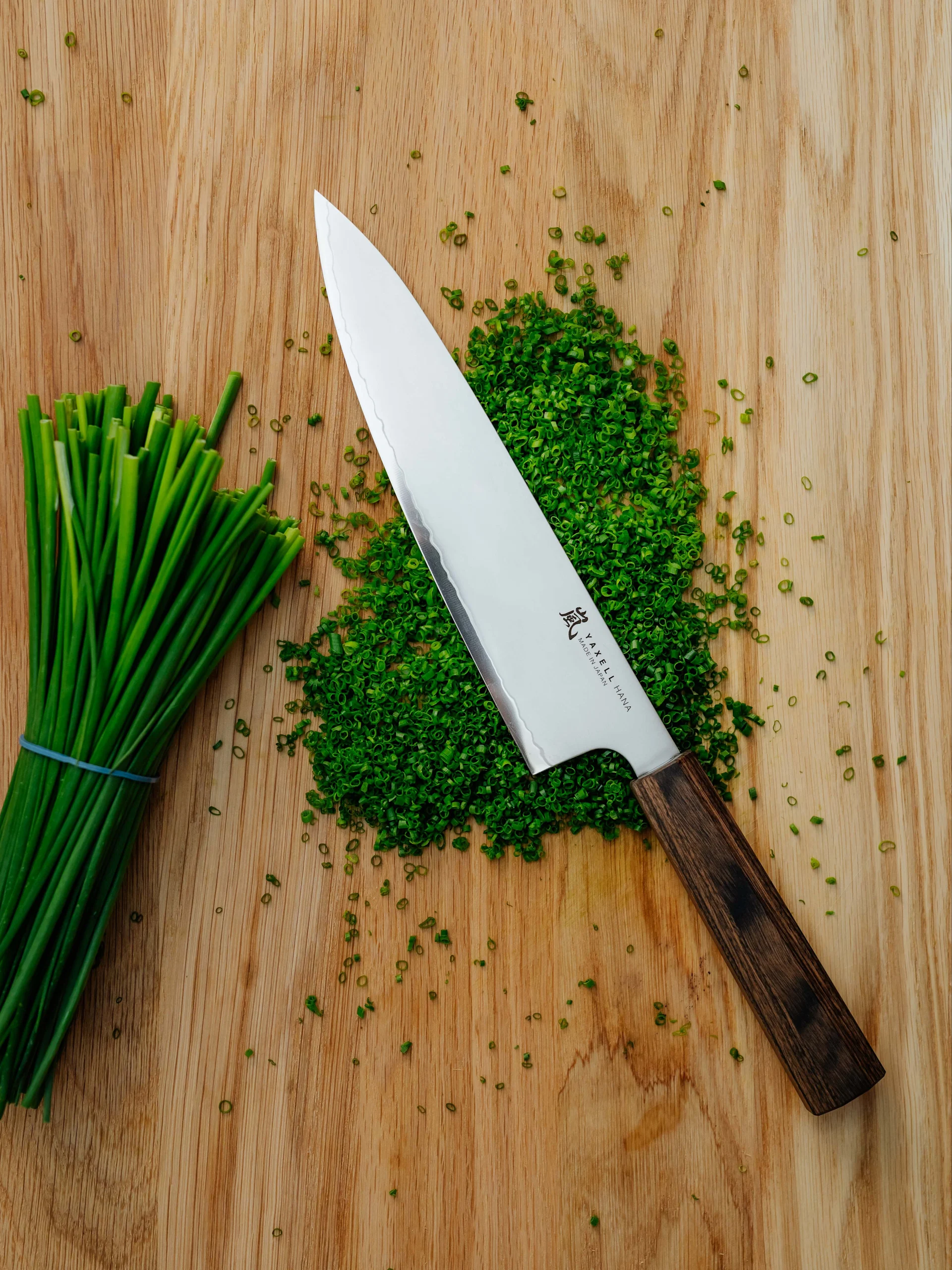 Kockkniv på en träskärbräda med en hög finhackad grön gräslök och en bunt hel gräslök.