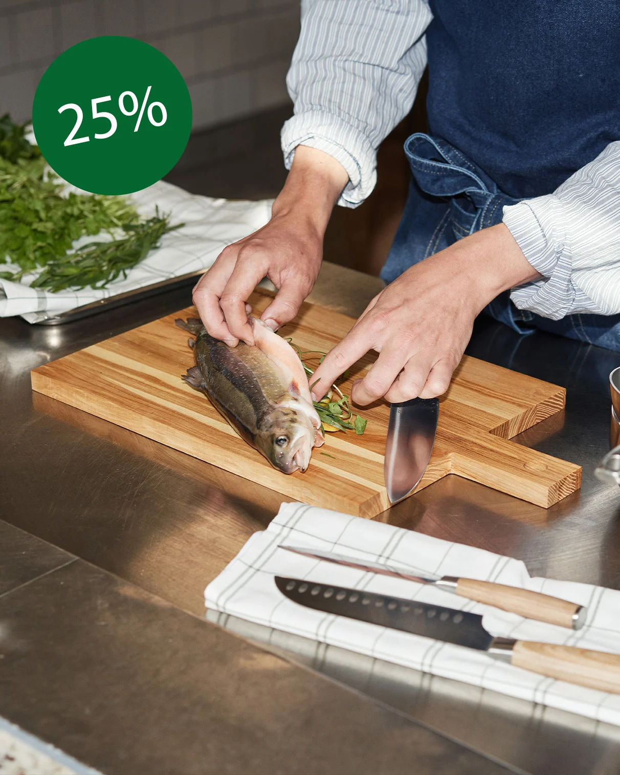 En person med denimförkläde rensar fisk på en träskärbräda. Bredvid ligger färska örter och knivar. Upptill syns '25%'