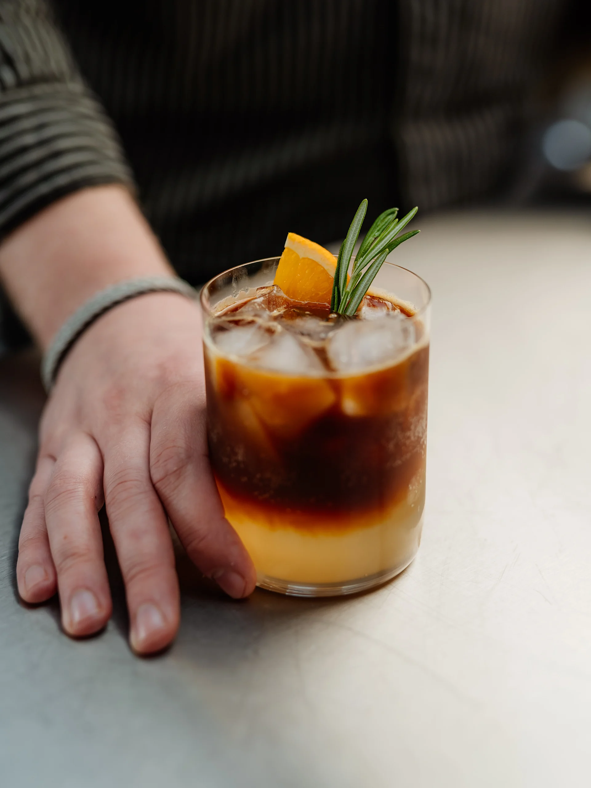 En hand håller en lagerisk kaffe med apelsinjuice, garnerad med en apelsinskiva och rosmarin.