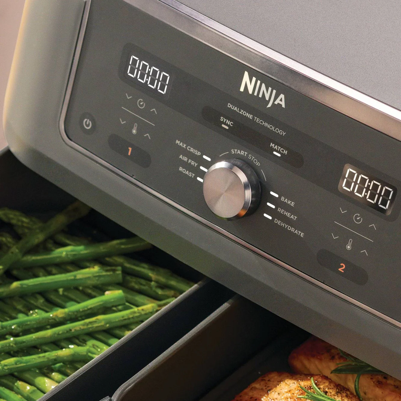 En Ninja airfryers kontrollpanel med två digitala displayer och en ratt, som visar grön sparris i den ena lådan och kyckling i den andra.
