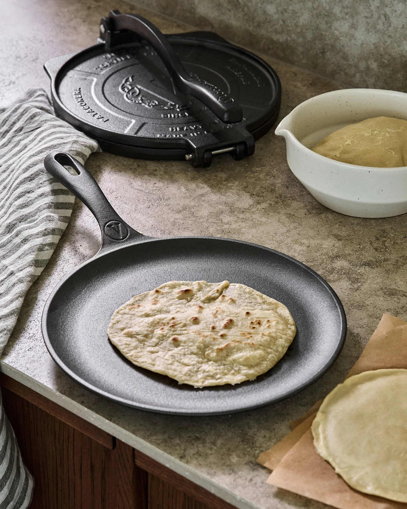Gjutjärnspanna med tunnbröd, bredvid en tortillapress och deg.