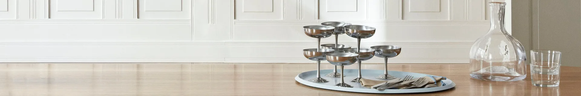 En ljusblå oval bricka med silverfärgade champagneglas, bestick och servett på ett ljust träbord bredvid en glaskaraff.